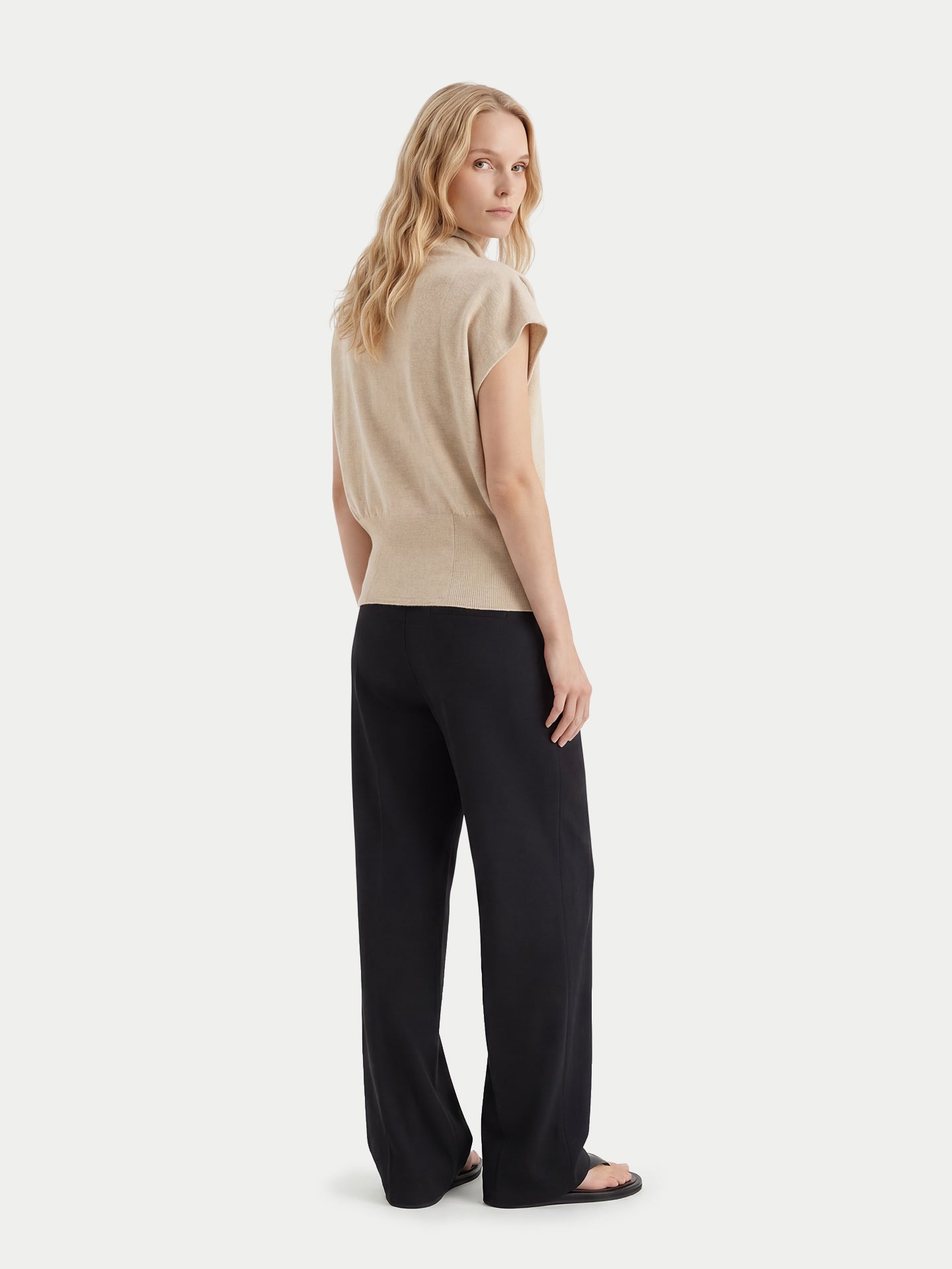 Vue de dos d'une femme debout portant un haut beige à manches courtes asymétrique, col côtelé à la taille et pantalon noir, sur fond gris clair. #color_beige