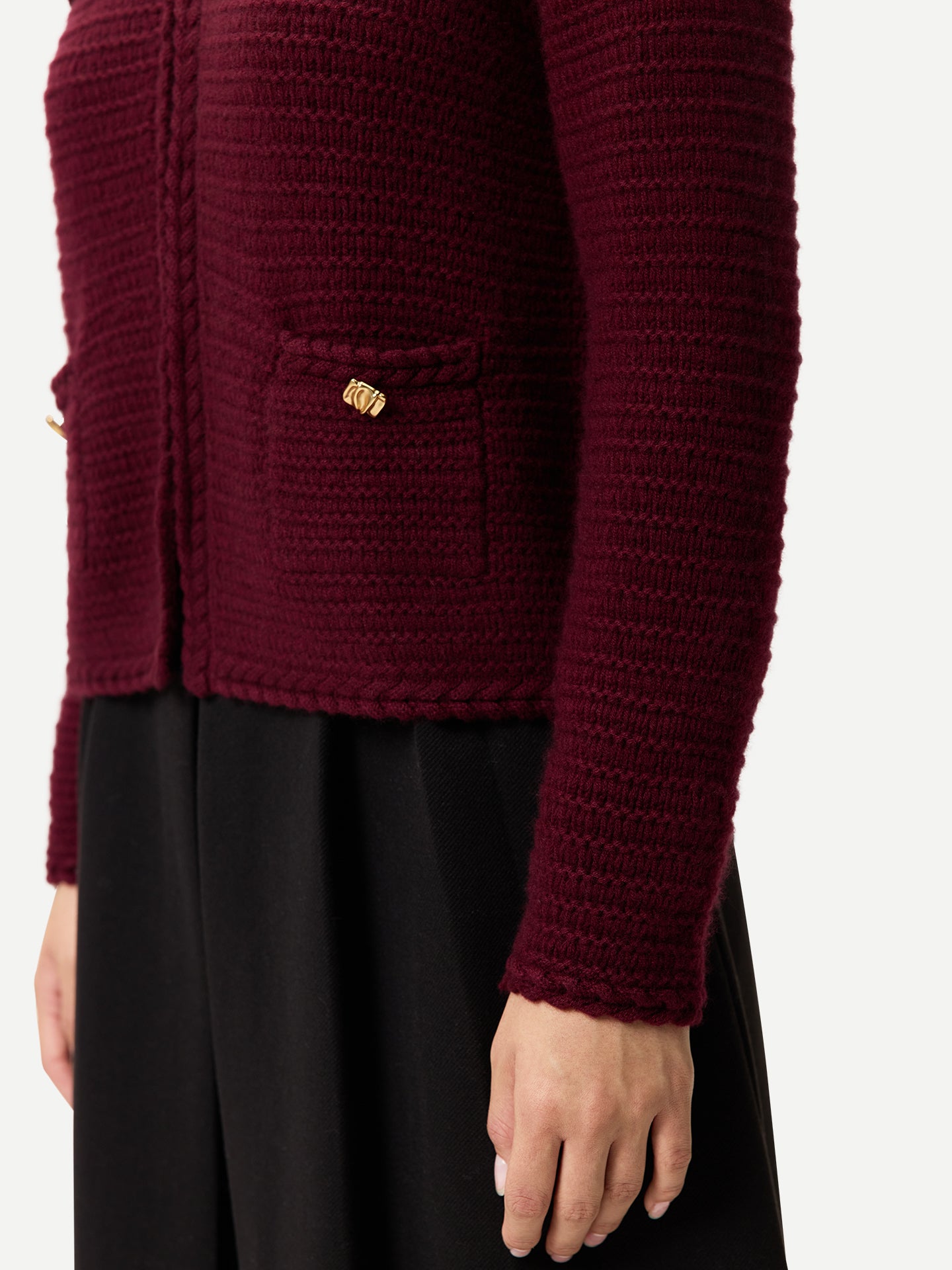 Gros plan de profil d'une femme portant un cardigan en maille bordeaux à boutons dorés sur les poches latérales et une jupe noire sur fond gris clair. #color_Zindanfil
