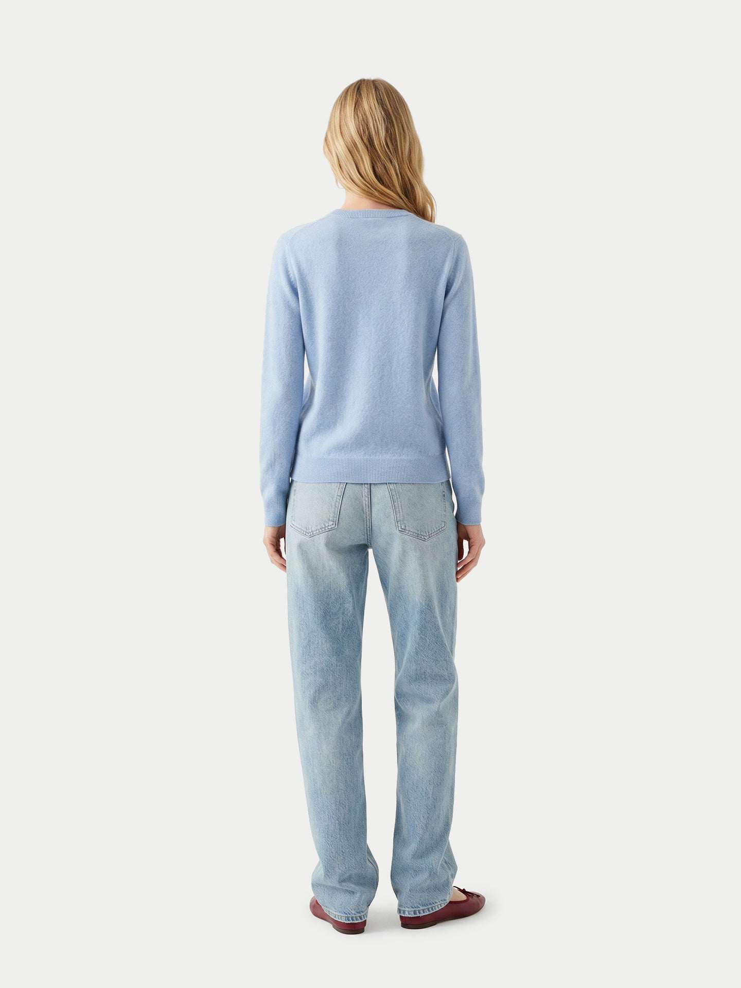 Vue de dos d'une femme portant un pull bleu clair, un jean bleu et des ballerines rouges sur un fond gris clair. #color_bleu-clair