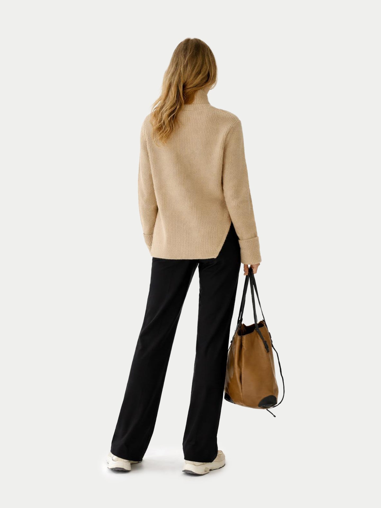 Vue de dos d'une femme portant un pull long beige côtelé à manches retroussées et fente latérale à la taille, et un pantalon noir sur fond gris clair. #color_beige