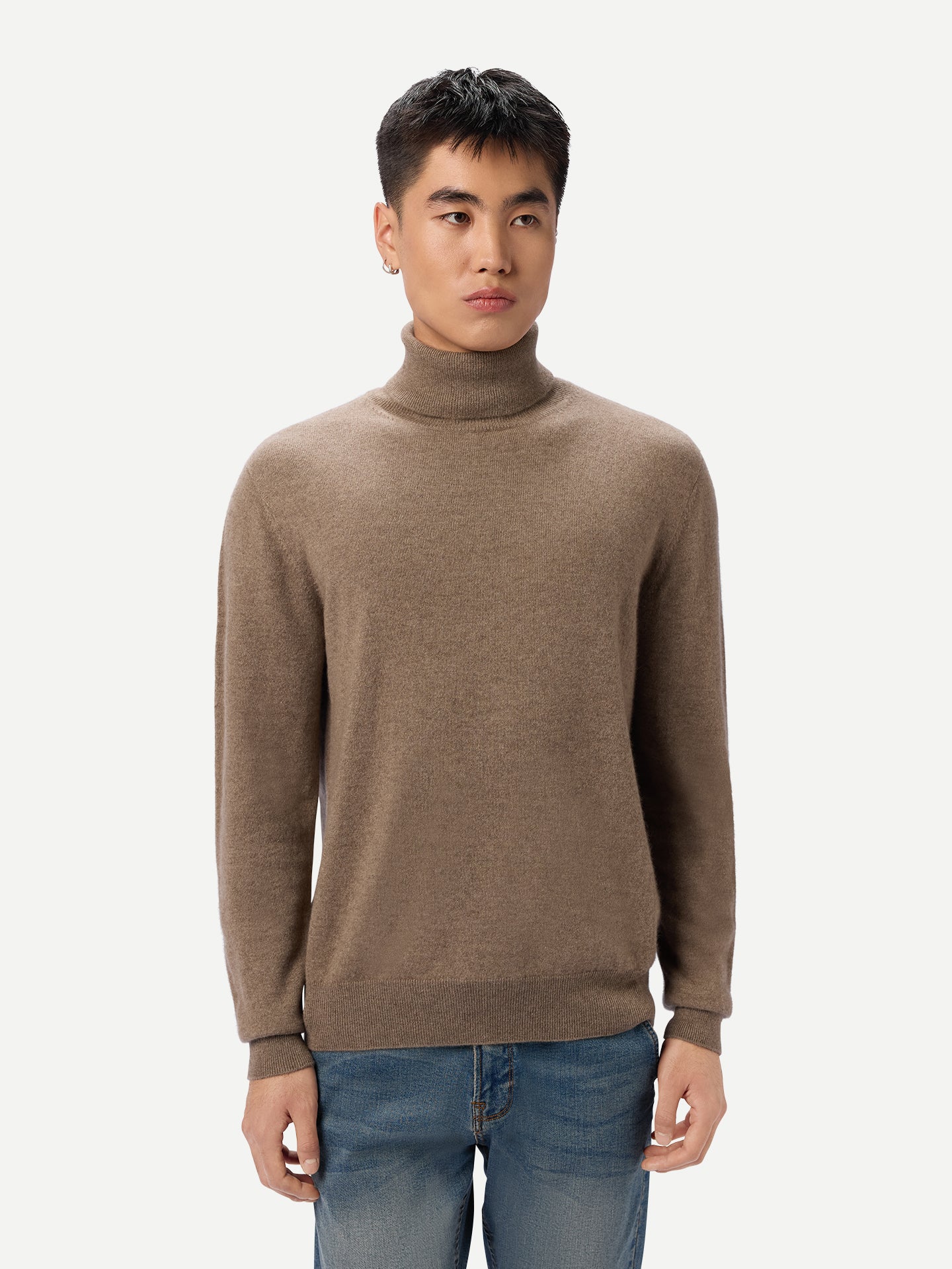 Gros plan de face d'un homme portant un pull à col roulé beige et un jean bleu sur fond gris clair. #color_taupe