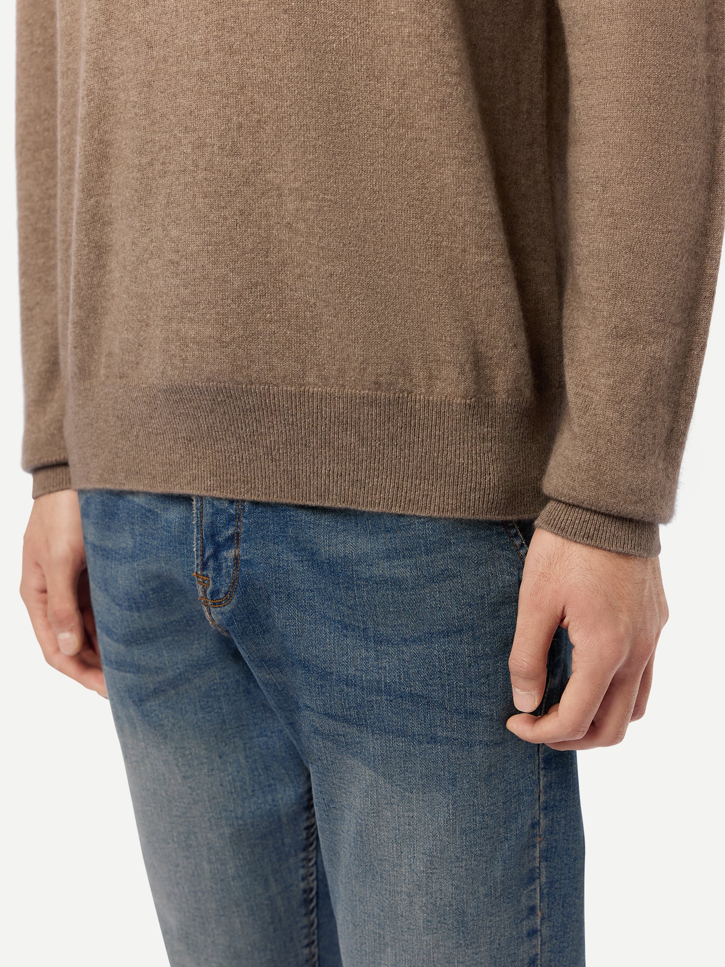 Gros plan sur le dessous d'un homme portant un pull beige à manches longues, poignets et ourlet côtelés, et un jean bleu, sur fond gris clair. #color_taupe