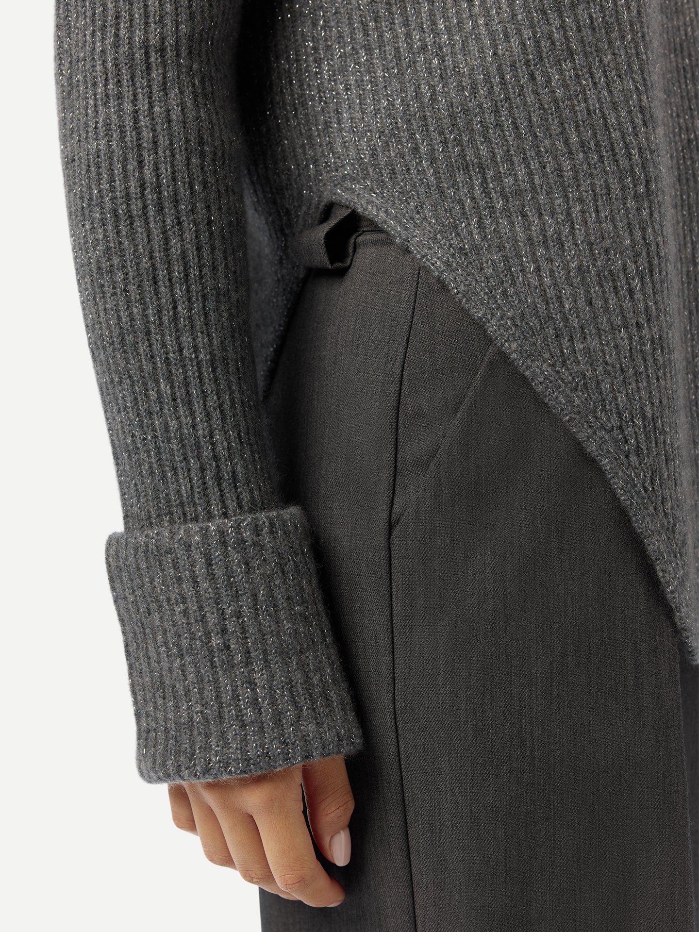 Gros plan sur une femme portant un pull long gris côtelé à manches retroussées et fente latérale à la taille, et un pantalon noir, sur fond gris clair. #color_gris-pierre