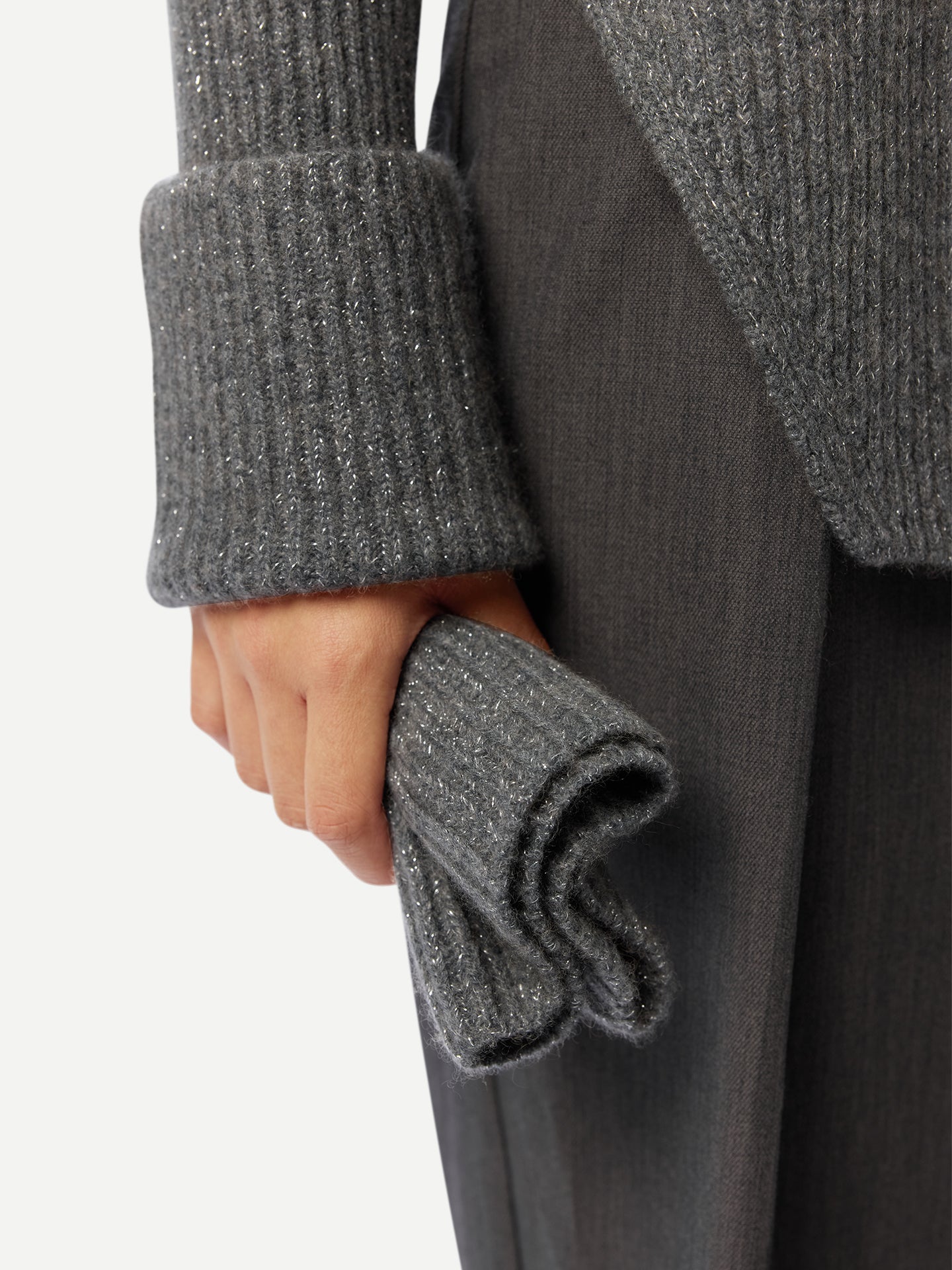 Gros plan sur une femme portant un pull long gris côtelé à manches retroussées et fente latérale à la taille, et un pantalon noir, tenant un cache-cou gris sur fond gris clair. #color_gris-pierre