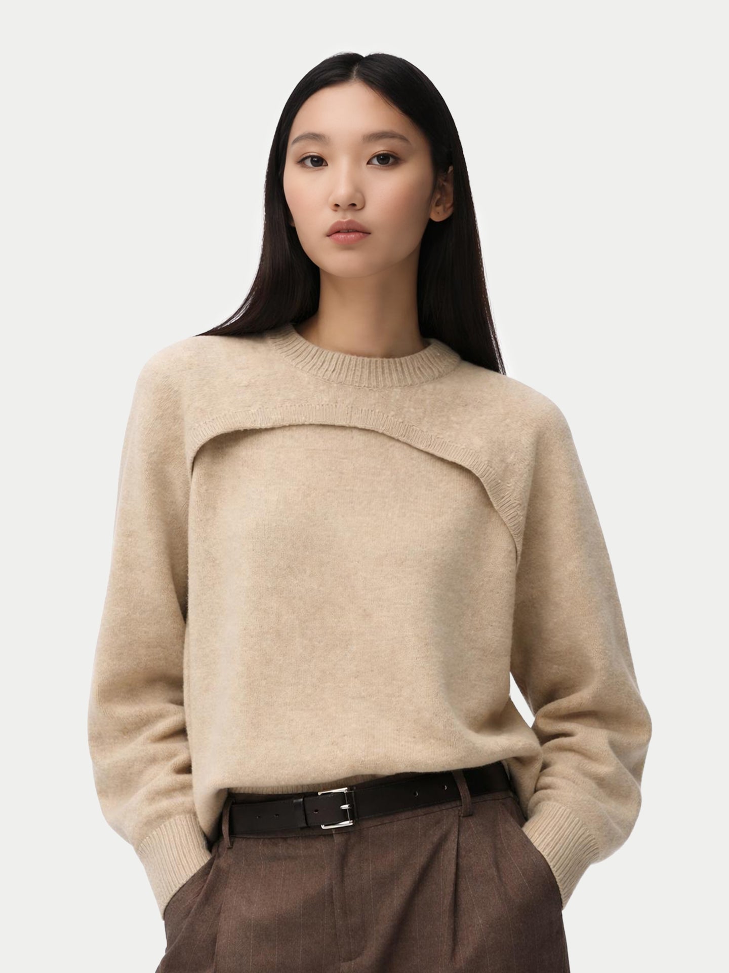 Gros plan de face d'une femme portant un pull beige en maille à motif architectural avec un empiècement sur la poitrine, associé à un pantalon en laine à fines rayures et une ceinture noire. #color_gris-chaud