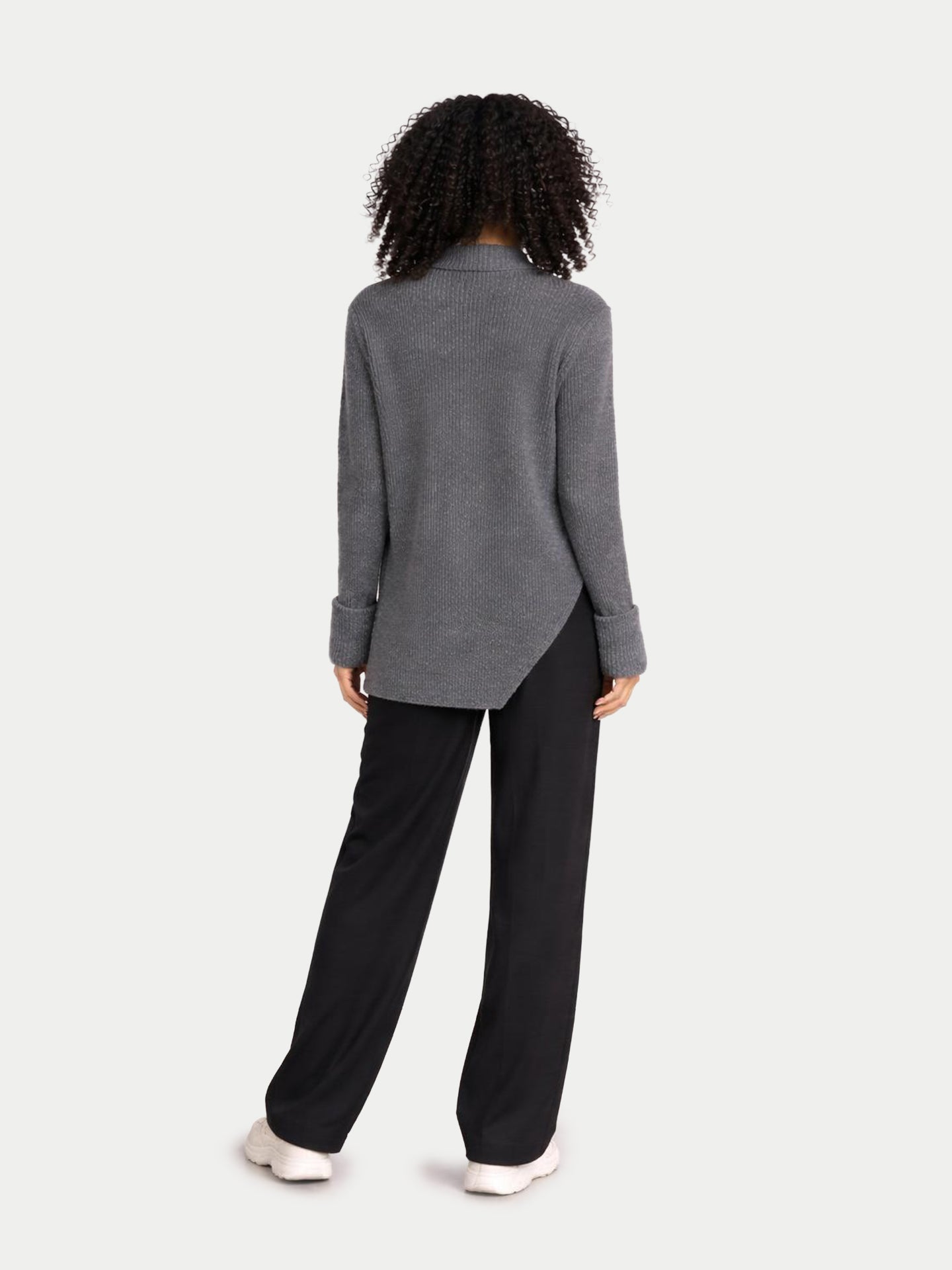 Vue de dos d'une femme portant un pull long gris côtelé à manches retroussées et fente latérale à la taille, et un pantalon noir sur fond gris clair. #color_gris-pierre