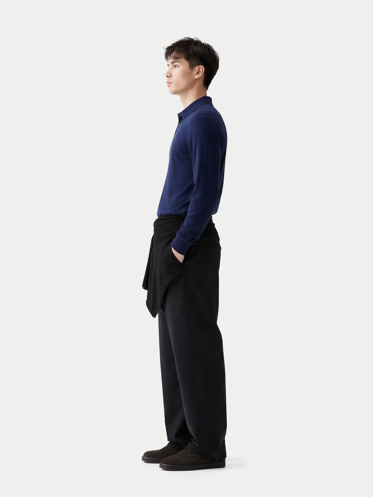 Vue de profil d'un homme portant un polo bleu marine à manches longues et un pantalon noir sur fond gris clair. #color_mood-indigo