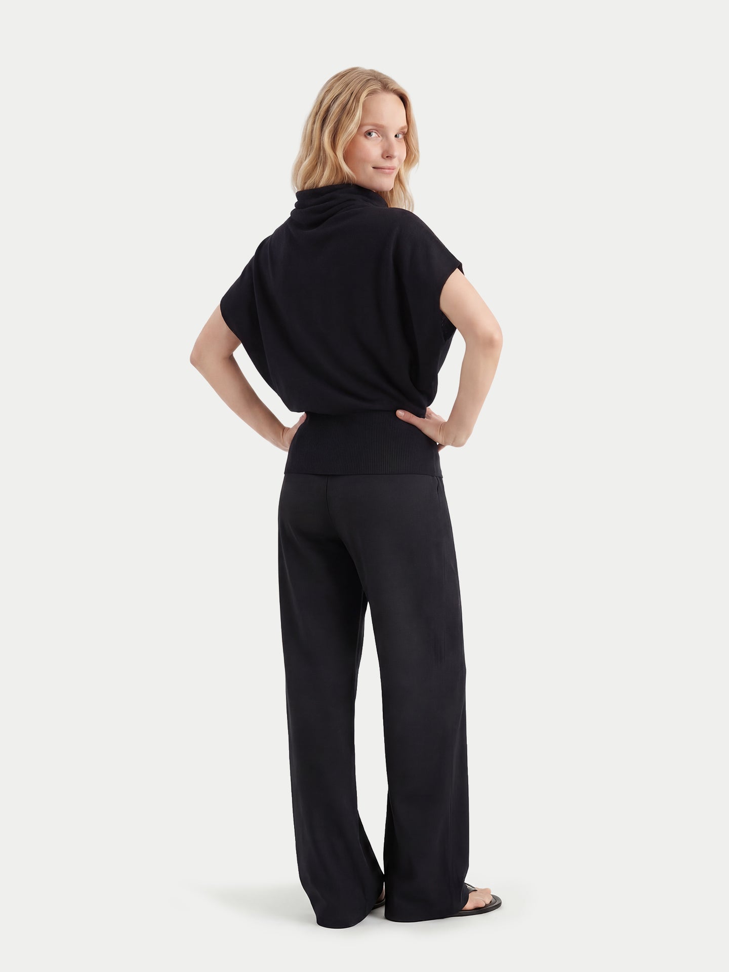 Vue de dos d'une femme portant un haut en cachemire noir à manches courtes, col asymétrique et taille côtelée, assorti à un pantalon large, sur un fond gris clair. #color_noir