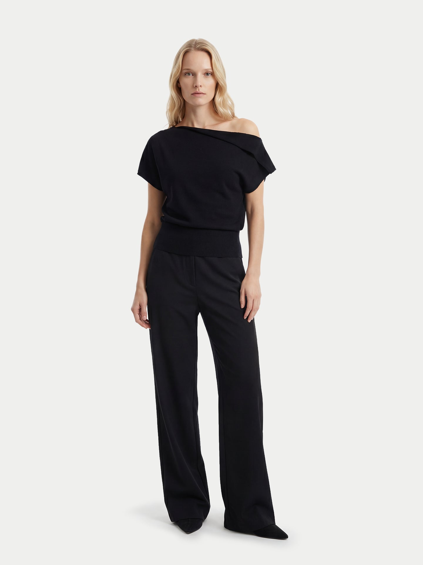 Vue de face d'une femme portant un haut en cachemire noir à manches courtes, col asymétrique et taille côtelée, assorti à un pantalon large, sur un fond gris clair. #color_noir