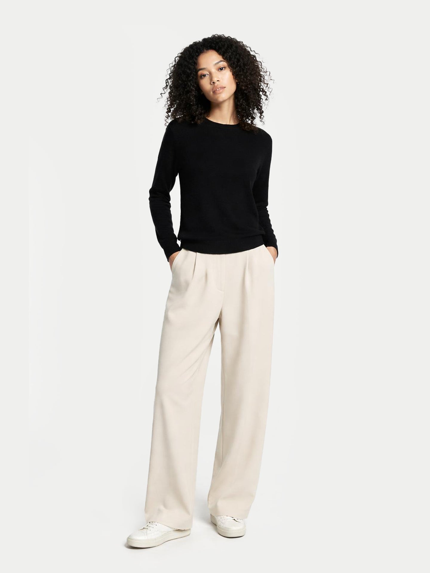 Vue de face d'une femme portant un pull noir à col rond, un pantalon blanc et des chaussures sur fond gris clair. #color_noir