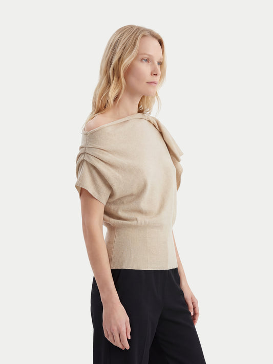 Gros plan de profil d'une femme portant un haut en cachemire beige à manches courtes, col asymétrique et taille côtelée, et un pantalon noir sur fond gris clair. #color_beige