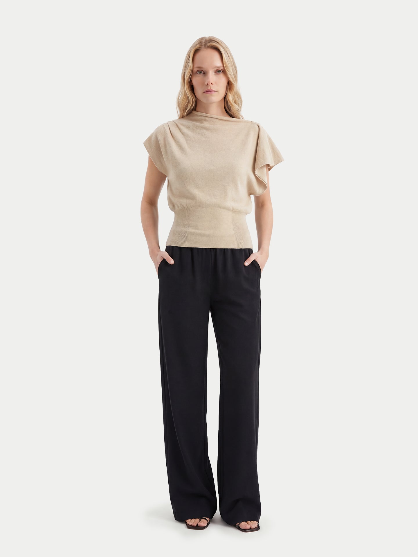 Vue de face d'une femme debout portant un haut beige à manches courtes asymétrique, col côtelé à la taille, en cachemire, et un pantalon noir sur fond gris clair. #color_beige