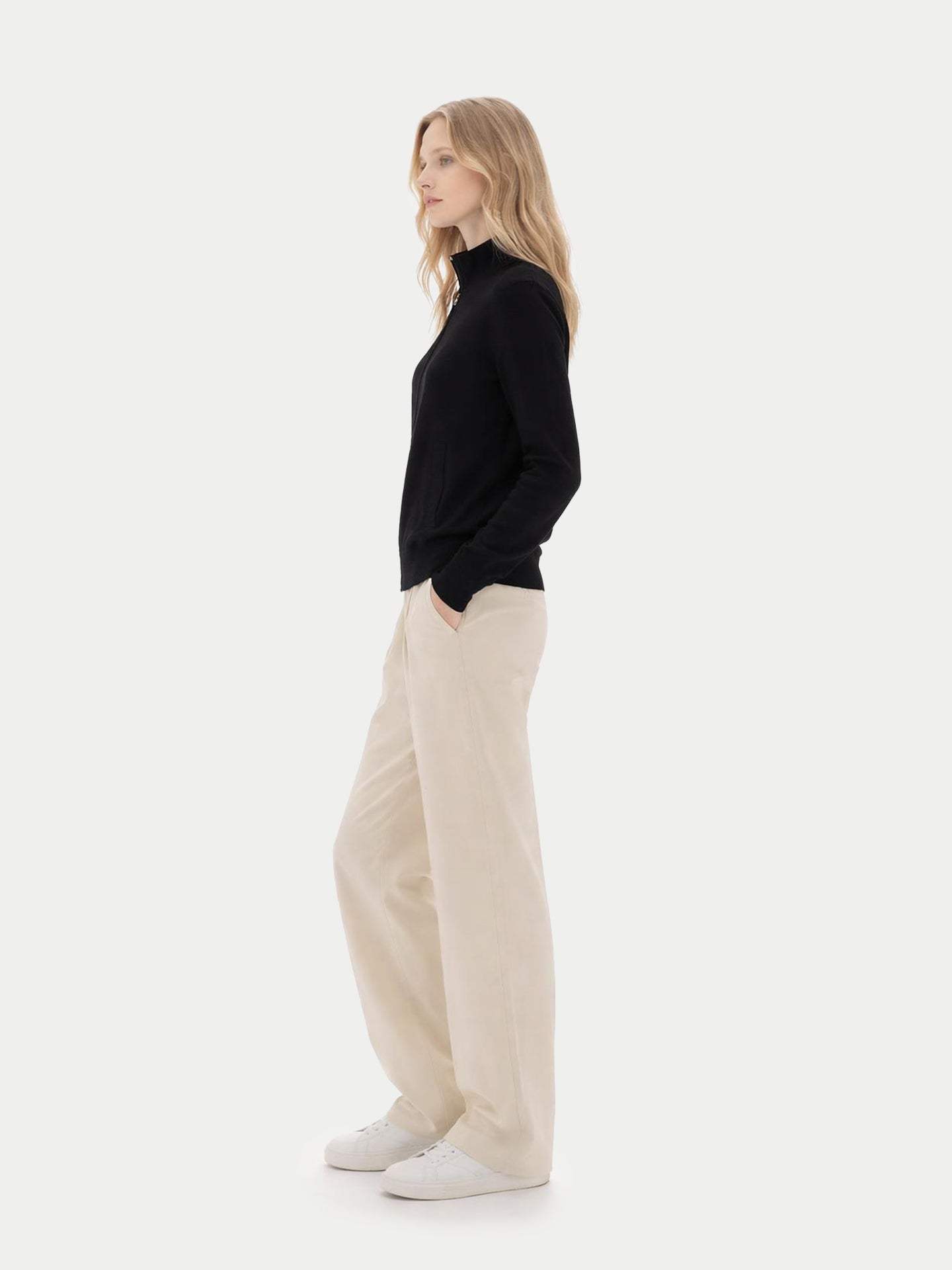 Vue de profil d'une femme portant un cardigan noir zippé à poches latérales et un pantalon blanc sur fond gris clair. #color_noir