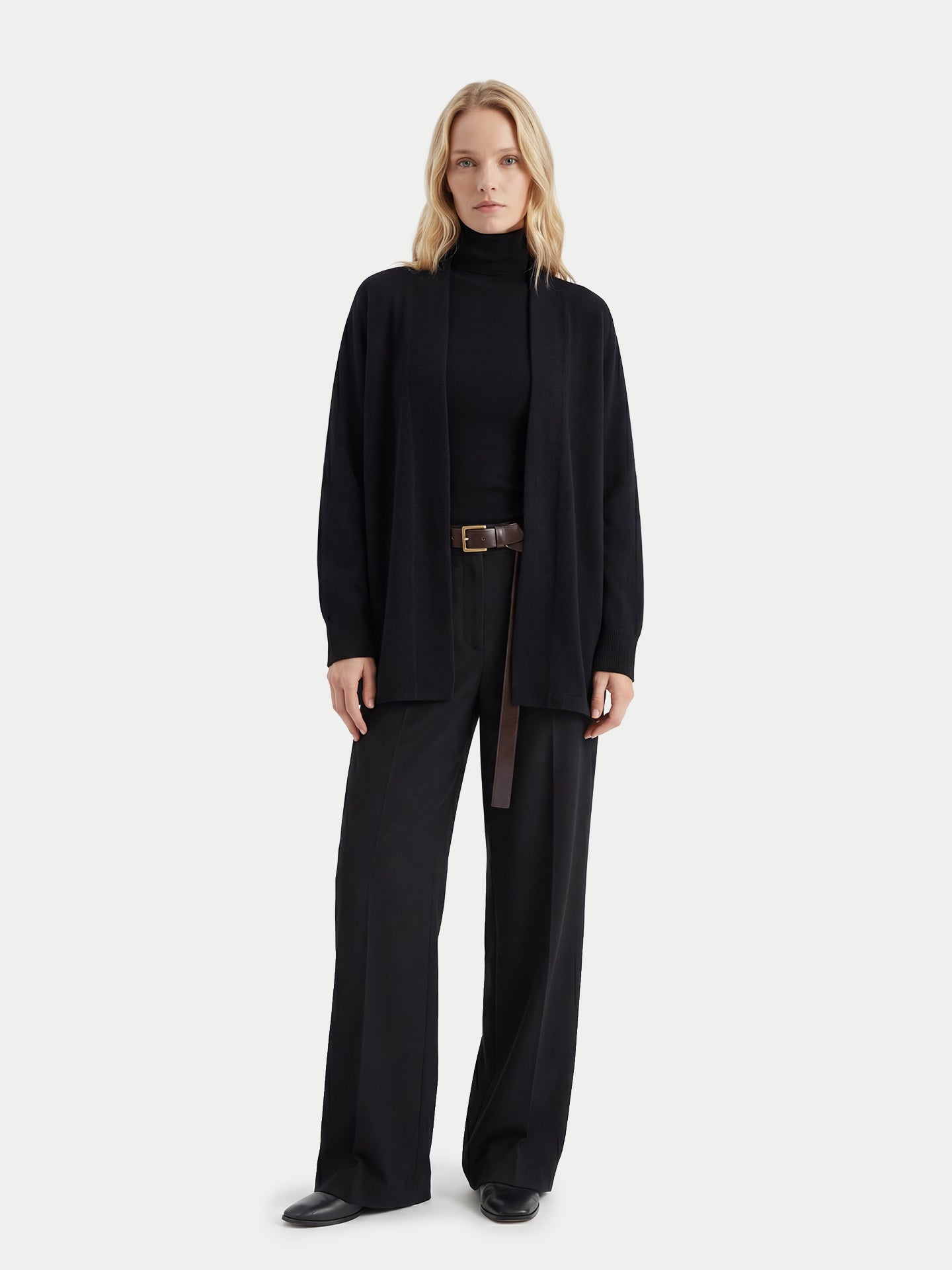 Vue de face d'une femme portant un cardigan noir ouvert sur un pull à col roulé noir et un pantalon assorti, sur fond gris clair. #color_noir