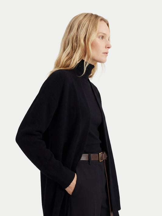 Gros plan de profil d'une femme portant un cardigan noir ouvert sur un pull à col roulé noir et un pantalon assorti, sur fond gris clair. #color_noir