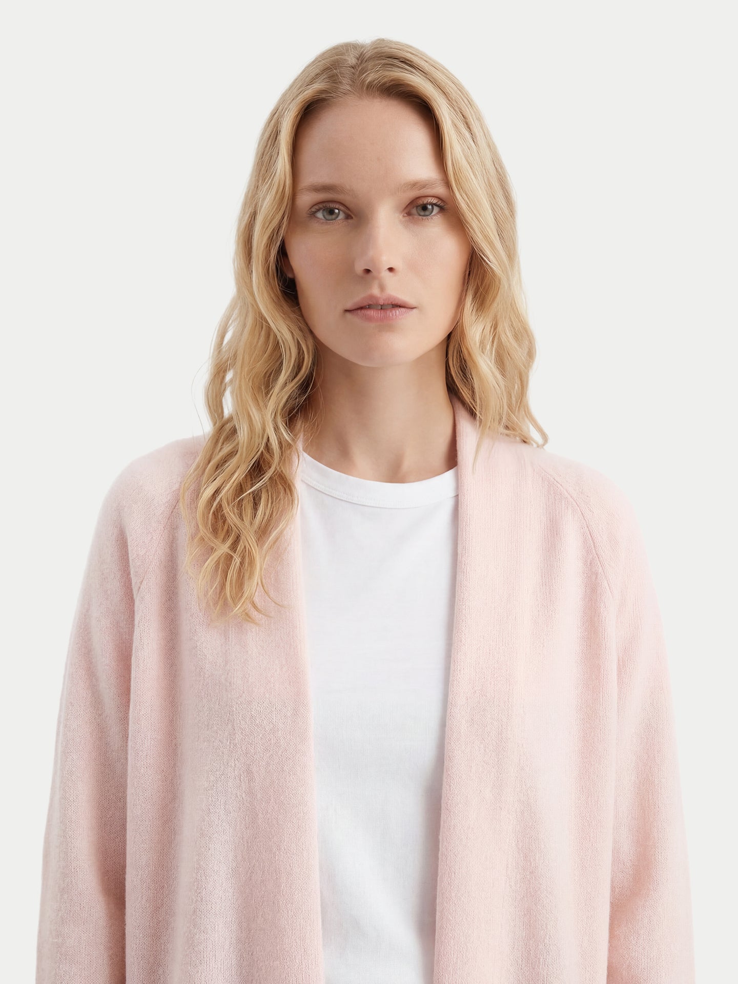 Gros plan de face d'une femme portant un cardigan rose pâle ouvert sur un t-shirt blanc, sur fond gris clair. #color_peach-blush