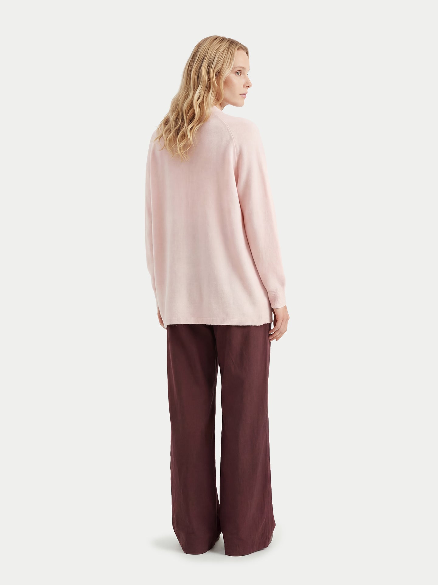 Vue de dos d'une femme portant un cardigan rose pâle et un pantalon bordeaux sur fond gris clair. #color_peach-blush
