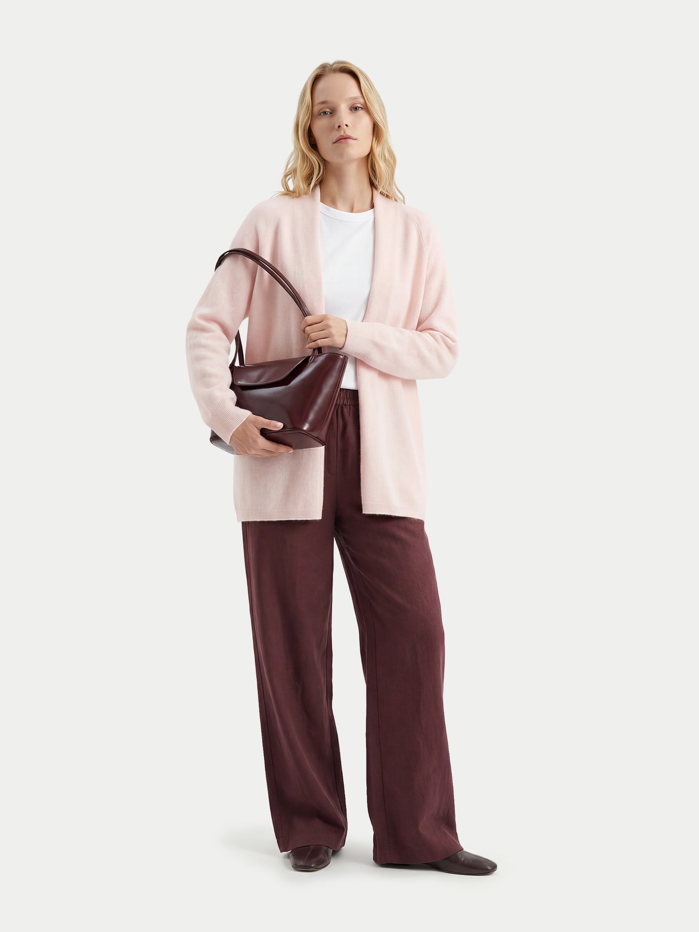 Vue de face d'une femme debout, portant un cardigan rose pâle ouvert sur un t-shirt blanc, tenant un sac à main bordeaux et un pantalon assorti, sur un fond gris clair. #color_peach-blush