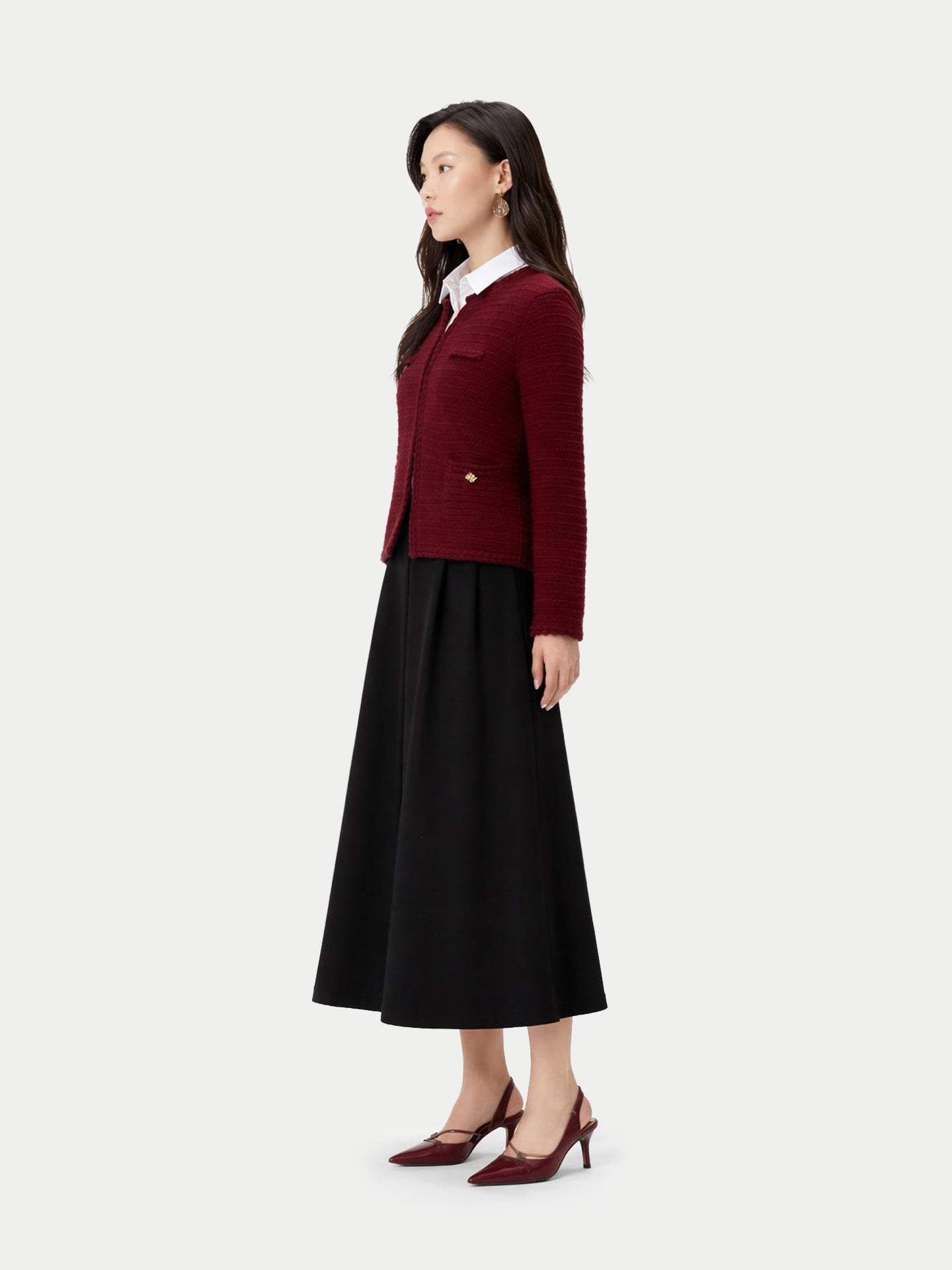 Vue de profil d'une femme portant un cardigan bordeaux en maille à col rond et boutons dorés sur les poches latérales, par-dessus une chemise blanche et une jupe noire, sur fond gris clair. #color_Zindanfil