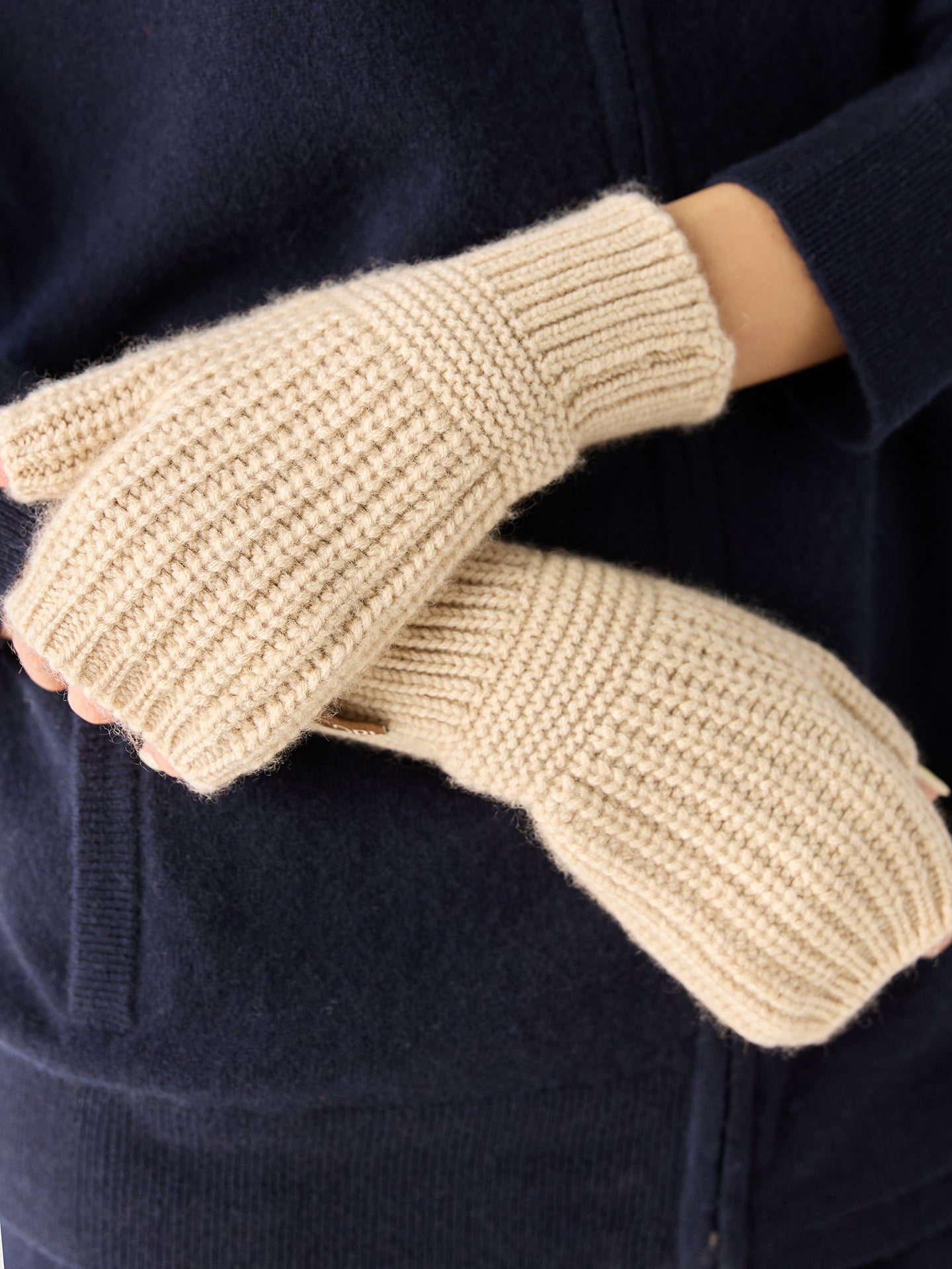 Gros plan sur une femme portant un ensemble bleu marine et des gants beiges sans doigts sur un fond gris clair. #color_beige