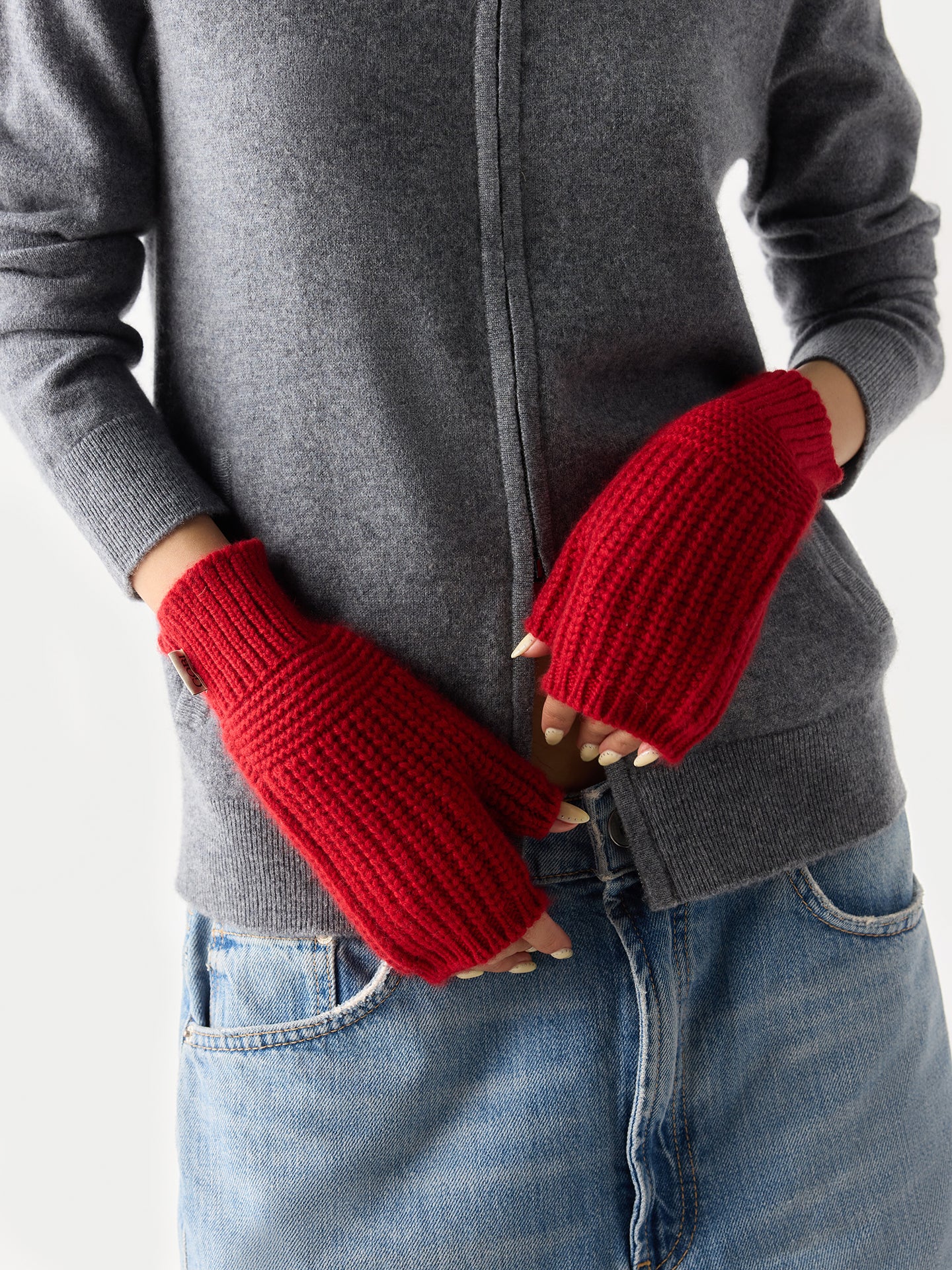 Gros plan de face d'une femme portant un pull gris à fermeture éclair, un jean bleu et des gants rouges sans doigts avec une petite étiquette de marque « GOBI » sur fond gris clair. #color_piment