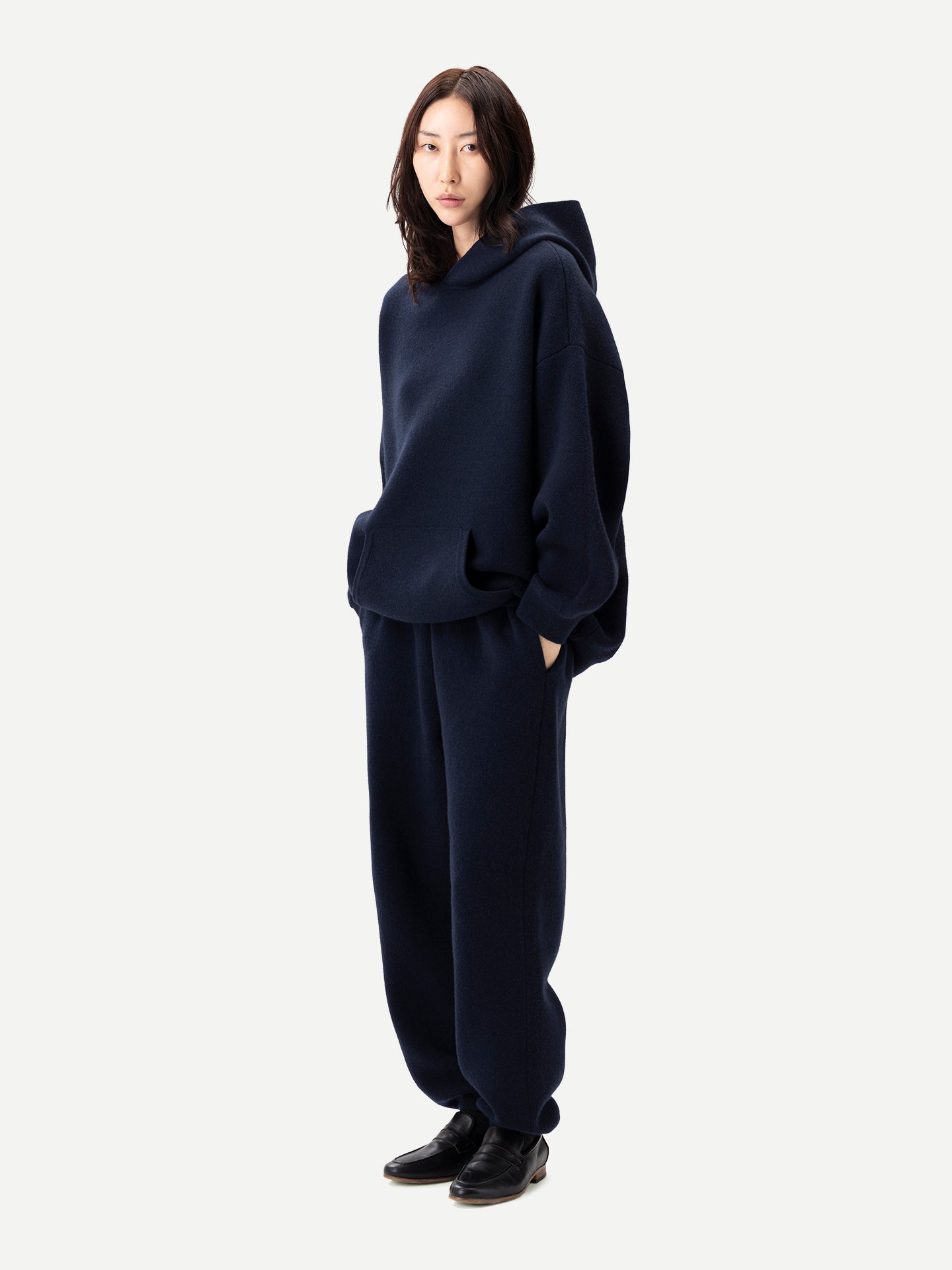 Vue de profil d'une femme debout, vêtue d'un sweat à capuche bleu marine assorti à une poche et d'un pantalon de jogging, sur fond gris clair. #color_india-ink