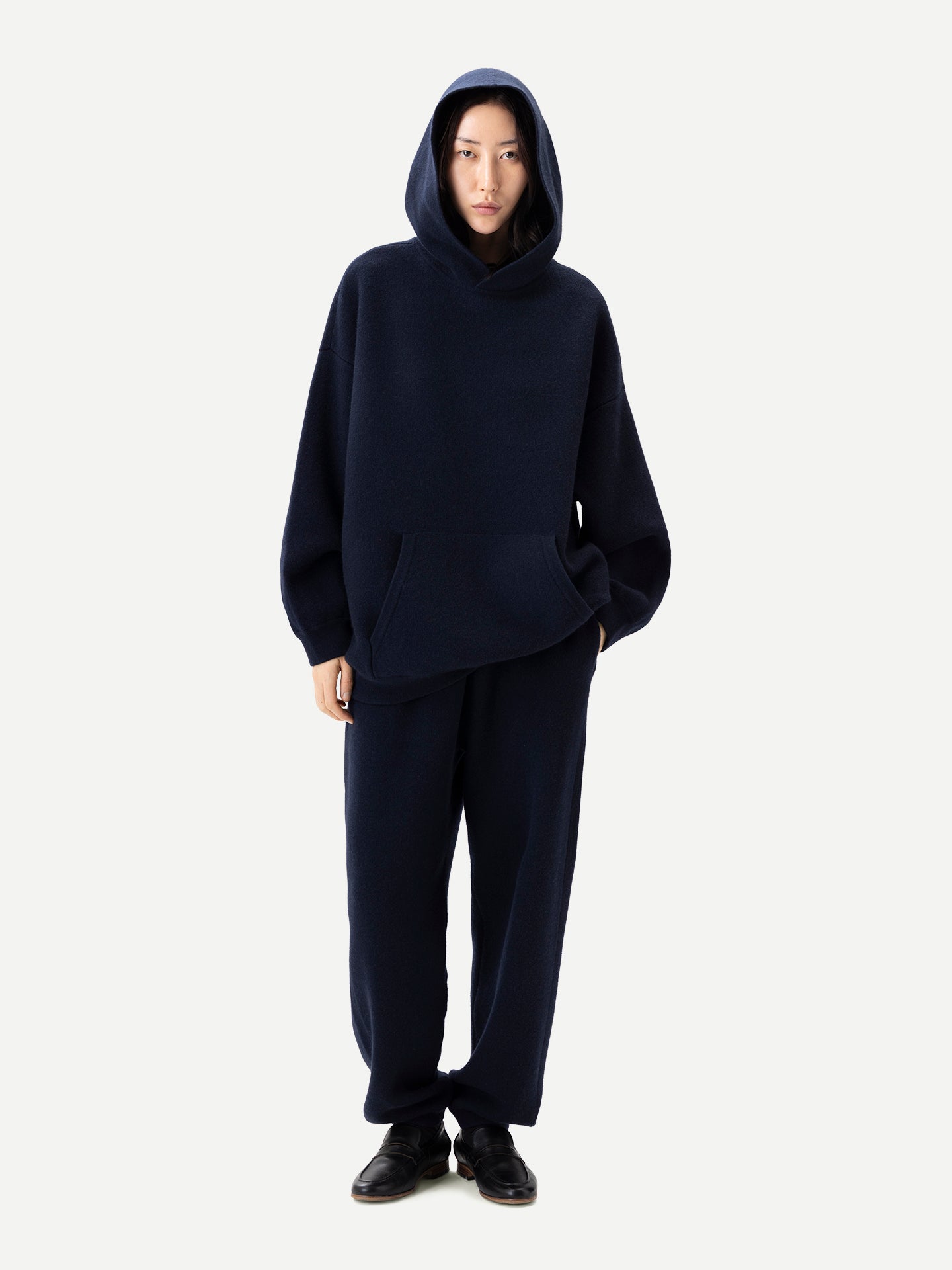 Vue de face d'une femme debout, vêtue d'un sweat à capuche bleu marine assorti à une poche et d'un pantalon de jogging, sur fond gris clair. #color_india-ink
