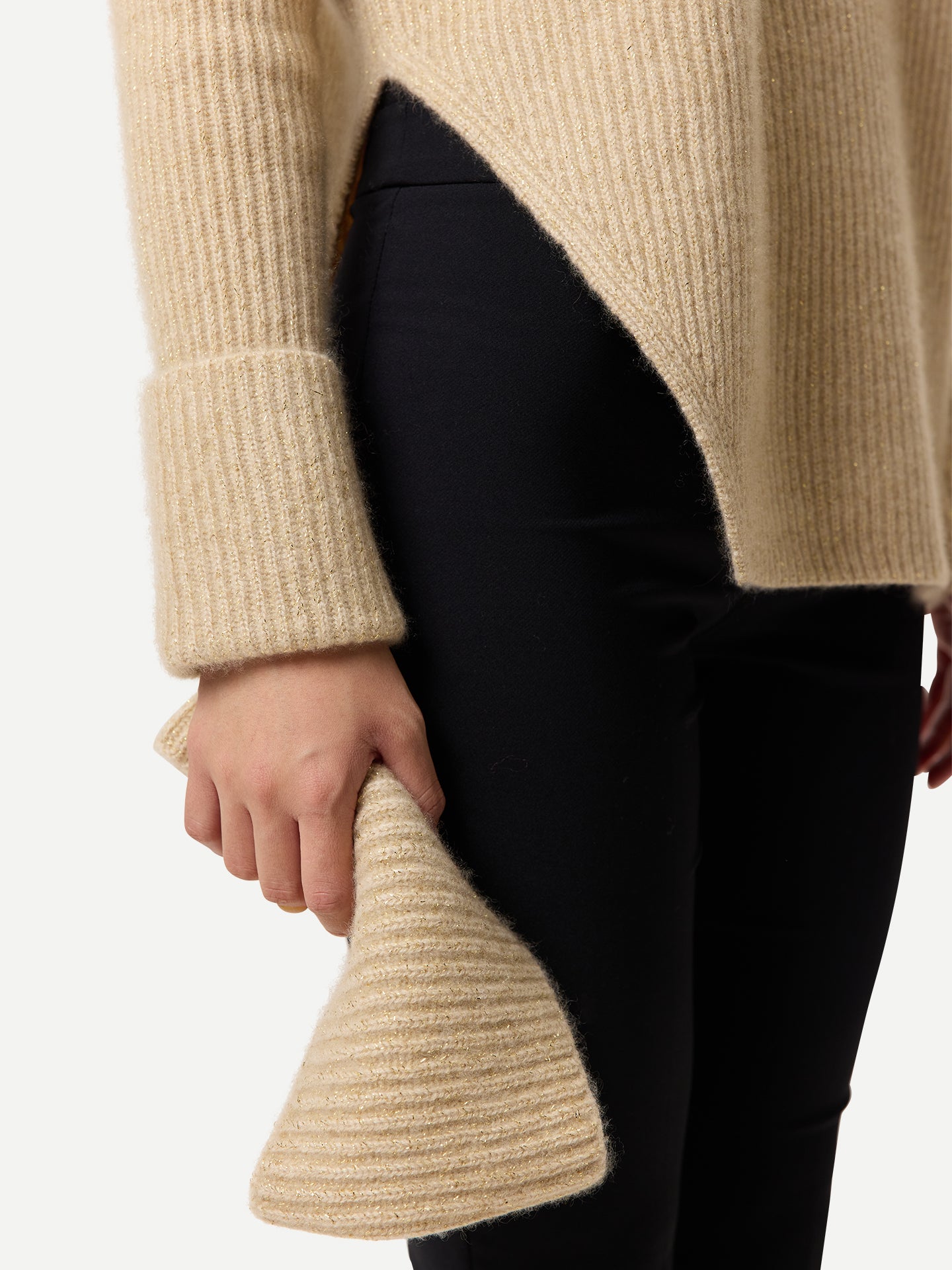 Gros plan sur une femme portant un pull long beige côtelé à manches retroussées et fente latérale à la taille, et un pantalon noir, tenant un cache-cou beige sur fond gris clair. #color_beige