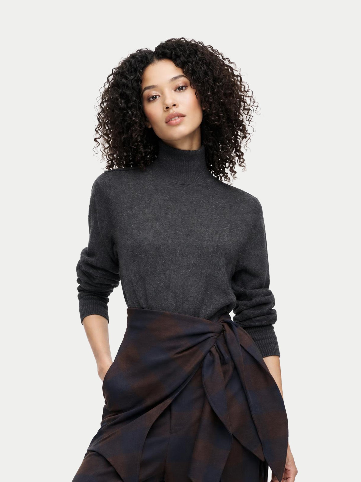 Un mannequin aux cheveux bouclés porte un pull à col roulé gris anthracite rentré dans un pantalon à carreaux marron foncé et marine orné d'un grand nœud décoratif à la taille. #color_gris-taupe