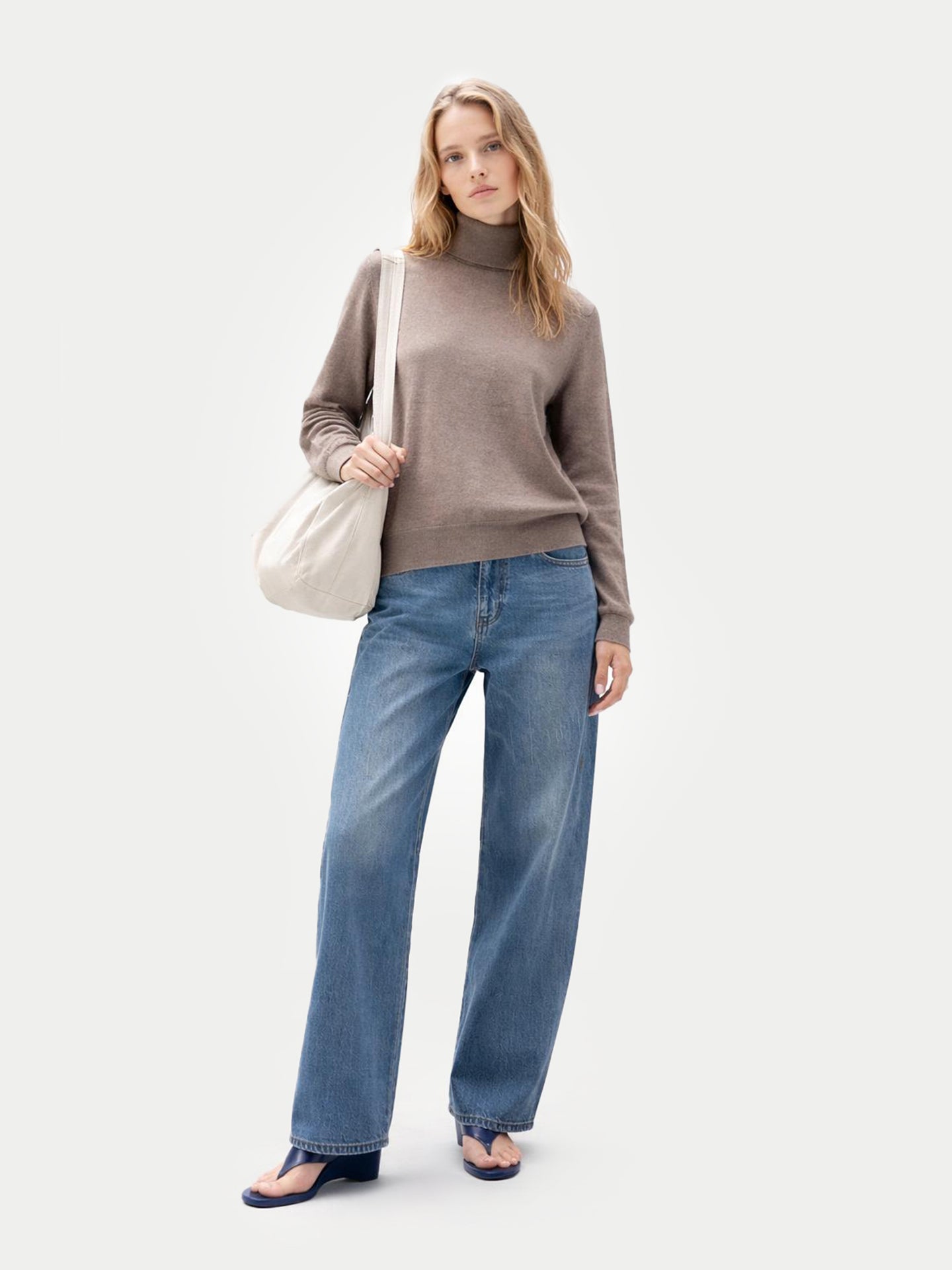 Vue de face d'une femme portant un pull à col roulé marron, un jean bleu et tenant un sac cabas blanc sur un fond gris clair. #color_taupe