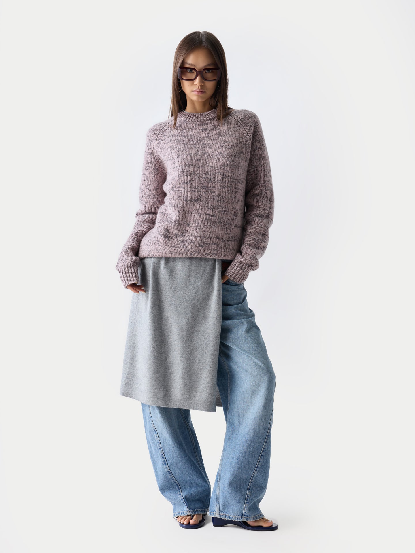 Vue de face d'une femme debout, portant des lunettes de soleil, un pull à col rond rose-gris sur une robe grise et un jean bleu, sur fond gris clair. #color_rose-ivoire