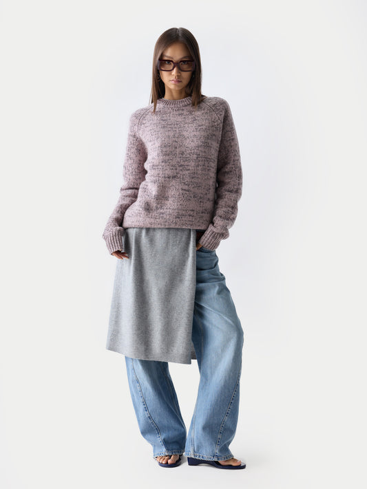 Vue de face d'une femme debout, portant des lunettes de soleil, un pull à col rond rose-gris sur une robe grise et un jean bleu, sur fond gris clair. #color_rose-ivoire