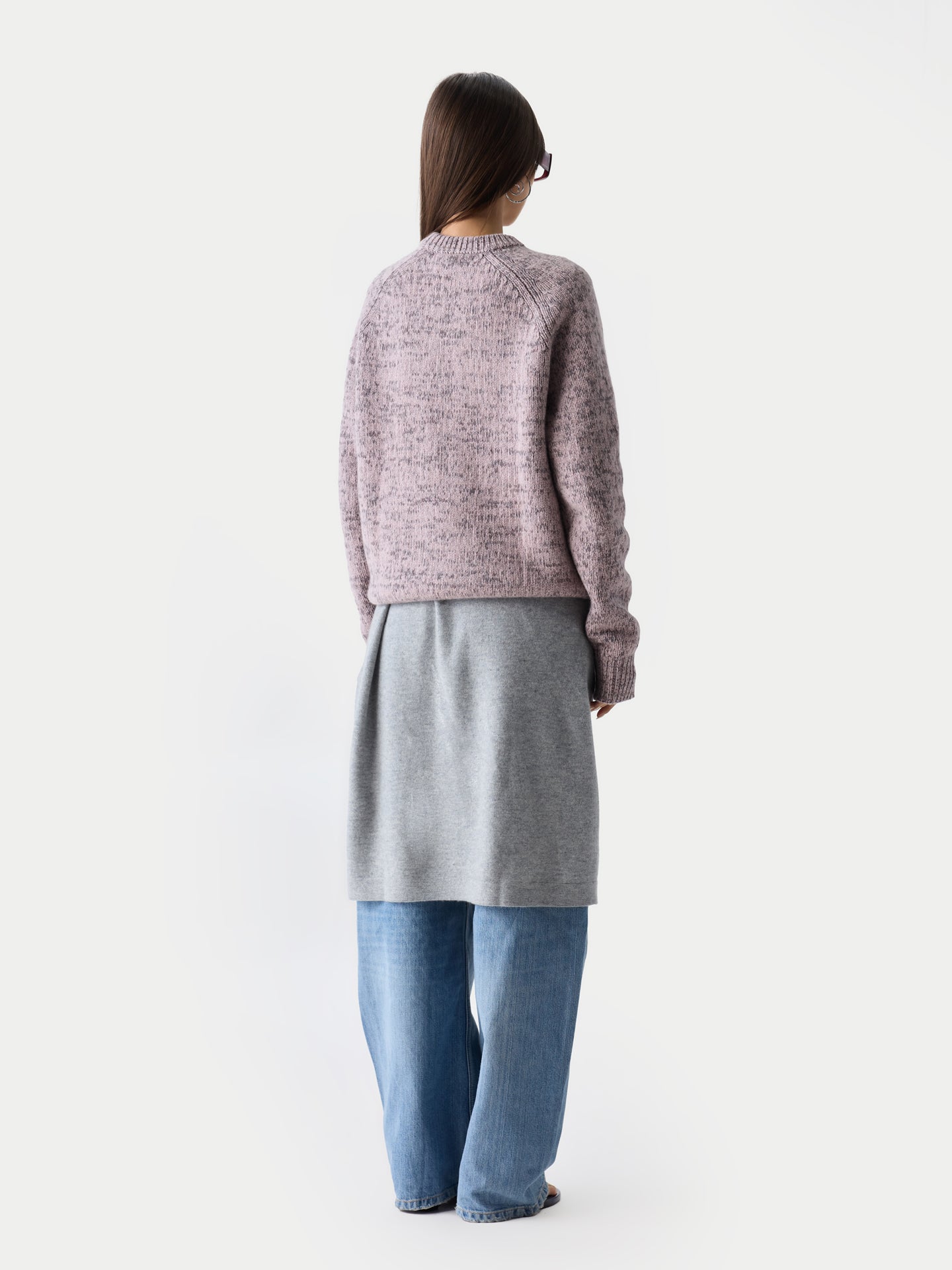 Vue de dos d'une femme debout, portant un pull ras du cou rose-gris sur une robe grise et un jean bleu, sur fond gris clair. #color_rose-ivoire
