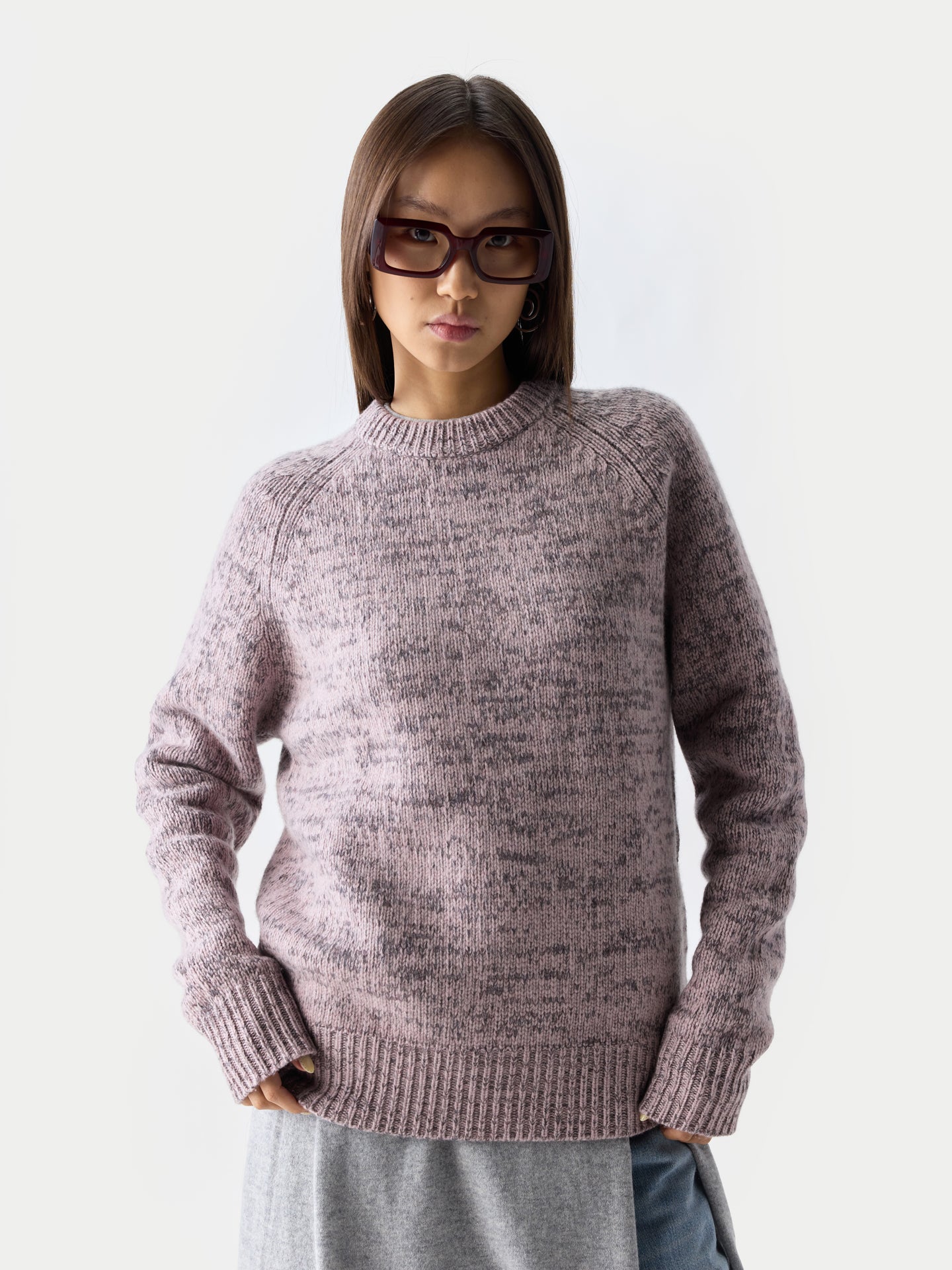 Gros plan de face d'une femme debout, portant des lunettes de soleil, un pull à col rond rose-gris sur une robe grise et un jean bleu, sur fond gris clair. #color_rose-ivoire