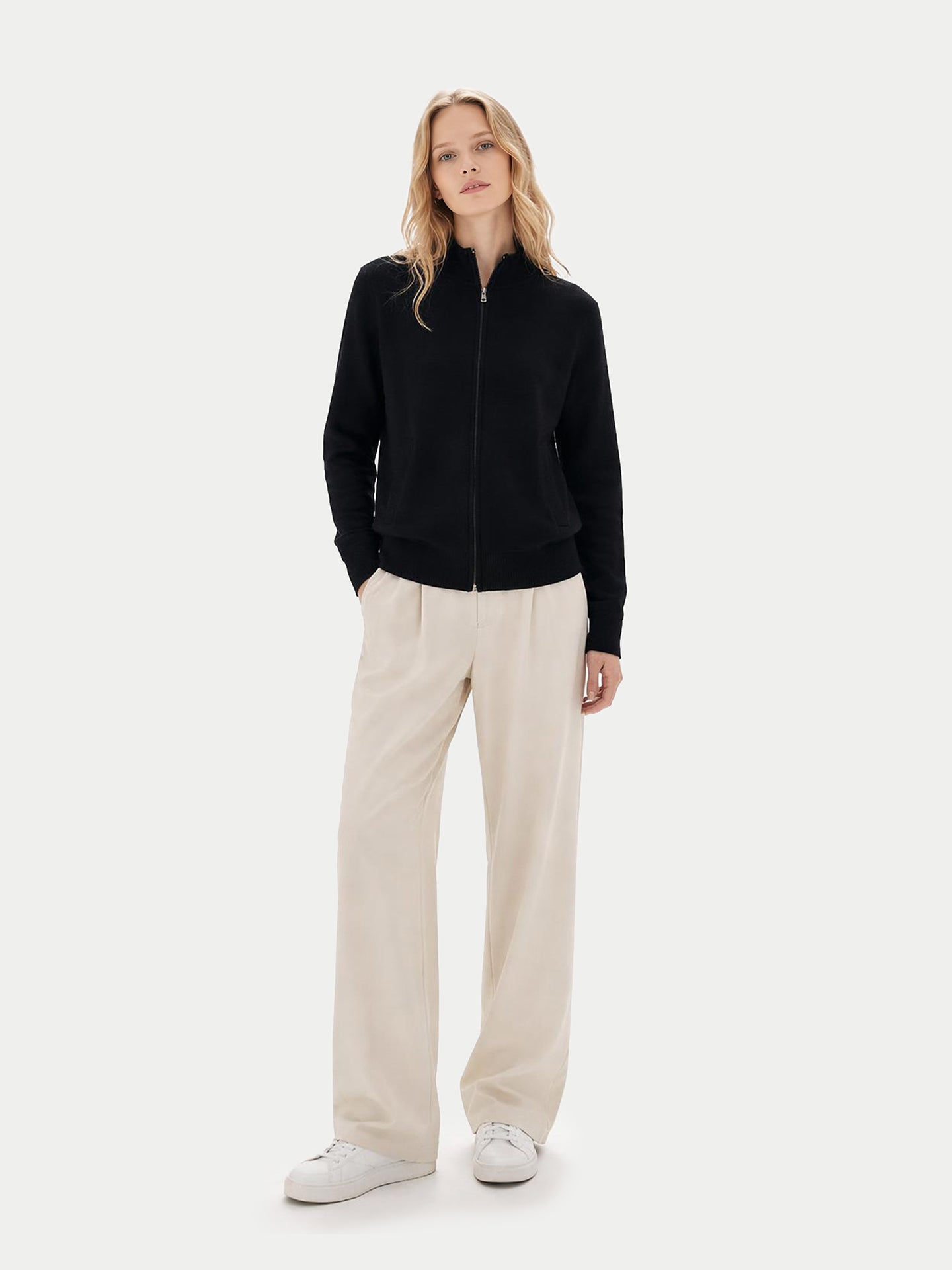 Vue de face d'une femme portant un cardigan noir zippé à poches latérales et un pantalon blanc sur fond gris clair. #color_noir