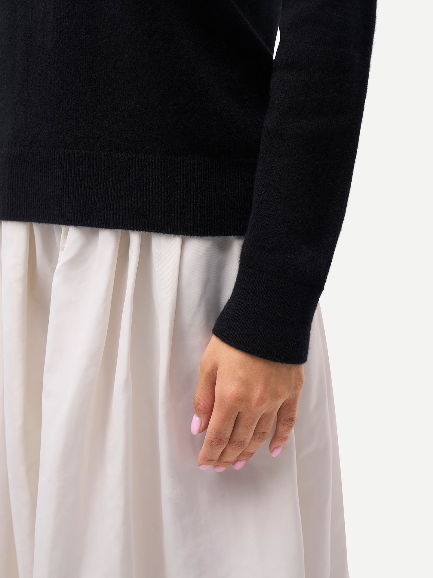 Gros plan sur le détail de la manche d'un pull à col roulé noir et d'une jupe blanche sur fond gris clair. #color_noir