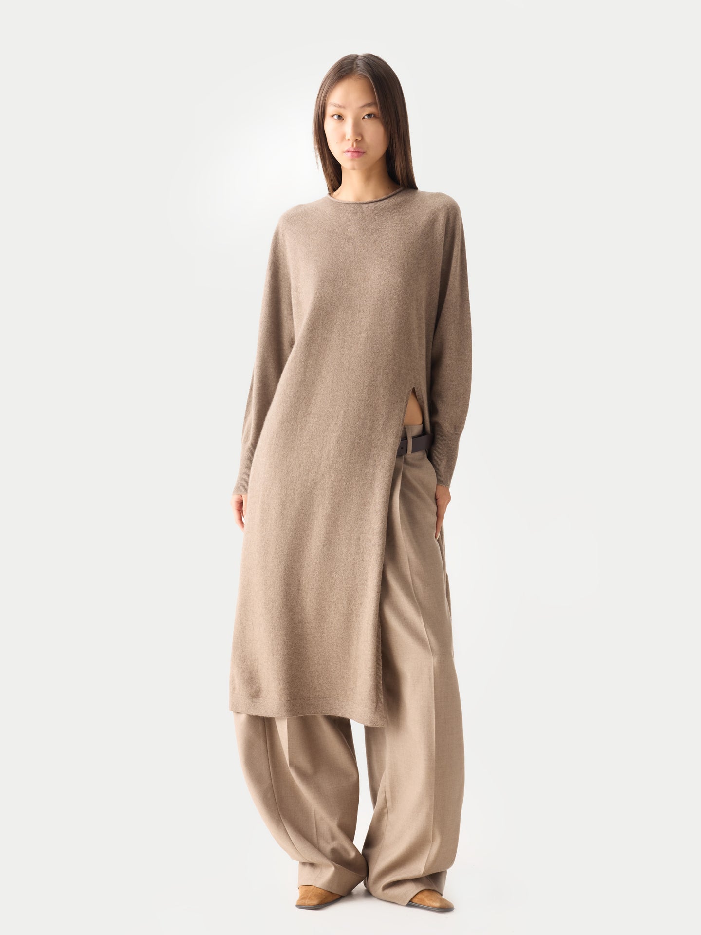 Vue de face d'une femme portant une robe longue beige à col rond et manches longues fendue sur le côté, sur un pantalon beige, sur fond gris clair. #color_taupe