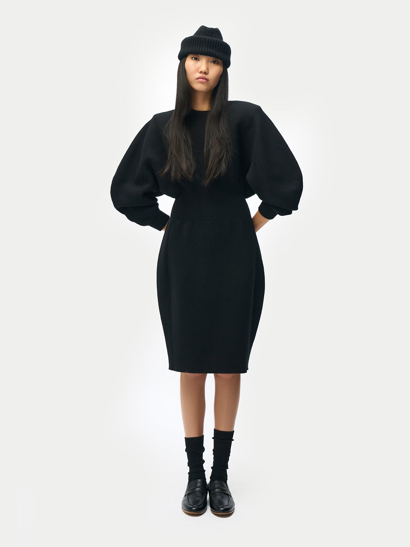 Vue de face d'une femme portant un chapeau noir et une robe midi noire à manches longues évasées, col rond et taille côtelée, sur fond gris clair. #color_noir