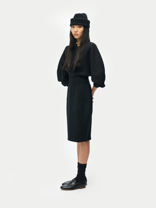 Vue de profil d'une femme portant un chapeau noir et une robe midi noire à manches longues évasées, col rond et taille côtelée, sur fond gris clair. #color_noir