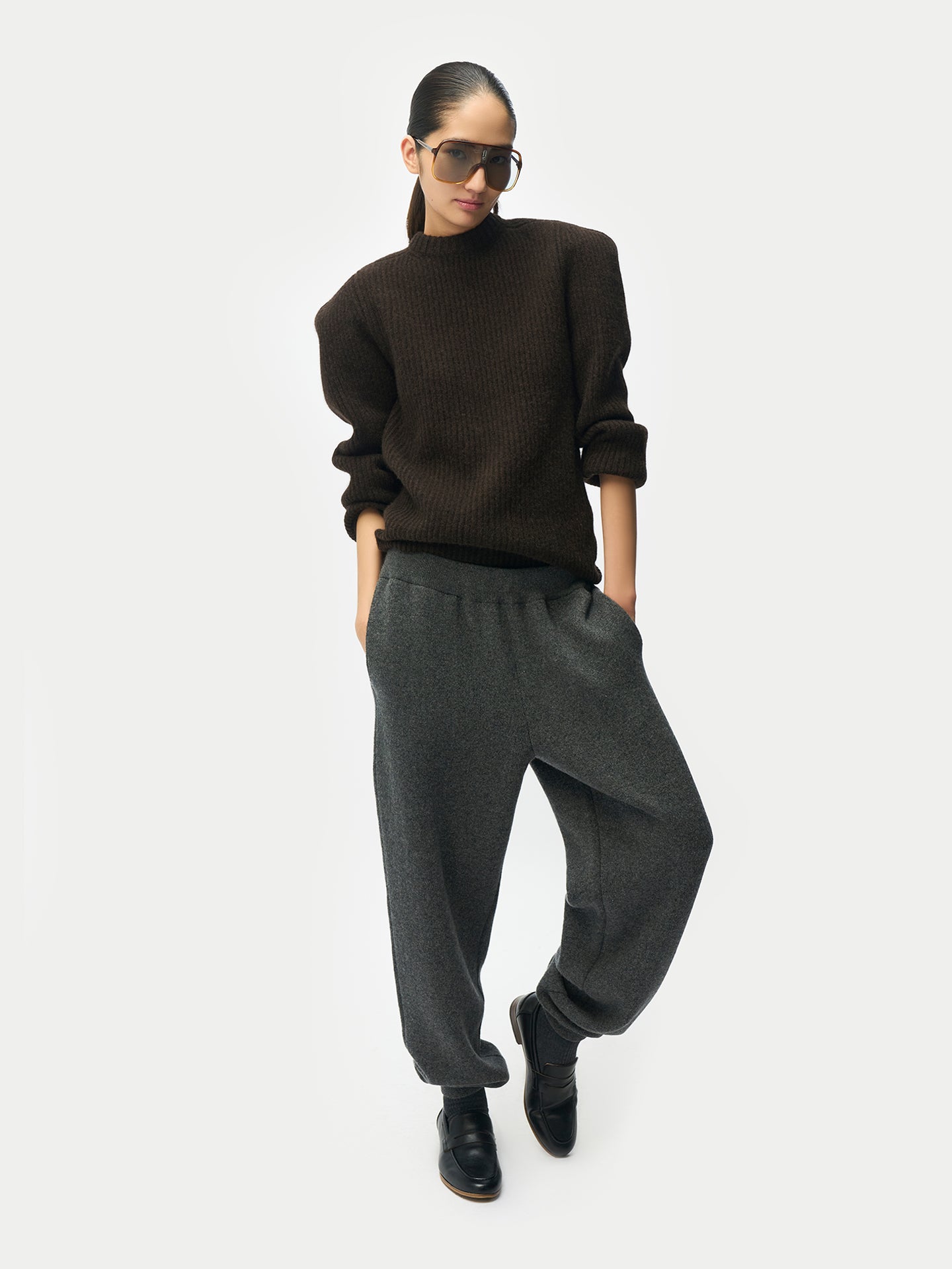 Vue de face d'une femme debout portant des lunettes de soleil, un pull côtelé marron et un pantalon de jogging gris, sur un fond gris clair. #color_mole