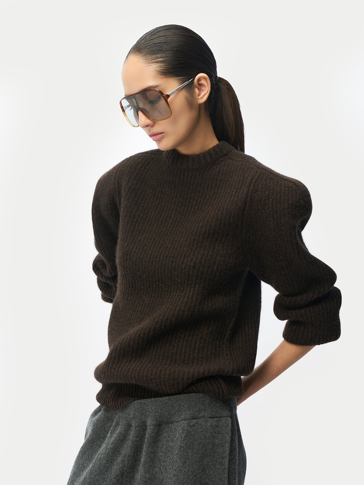 Gros plan de face d'une femme debout portant des lunettes de soleil, un pull côtelé marron et un pantalon gris sur fond gris clair. #color_mole