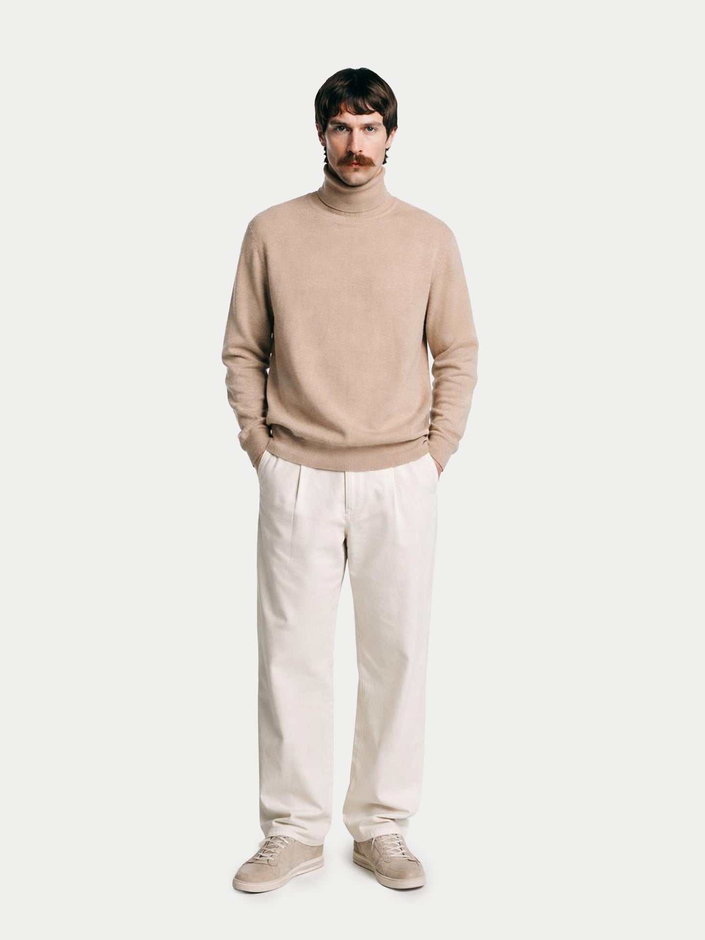 Vue de face d'un homme portant un pull à col roulé beige et un pantalon blanc sur fond gris clair. #color_beige