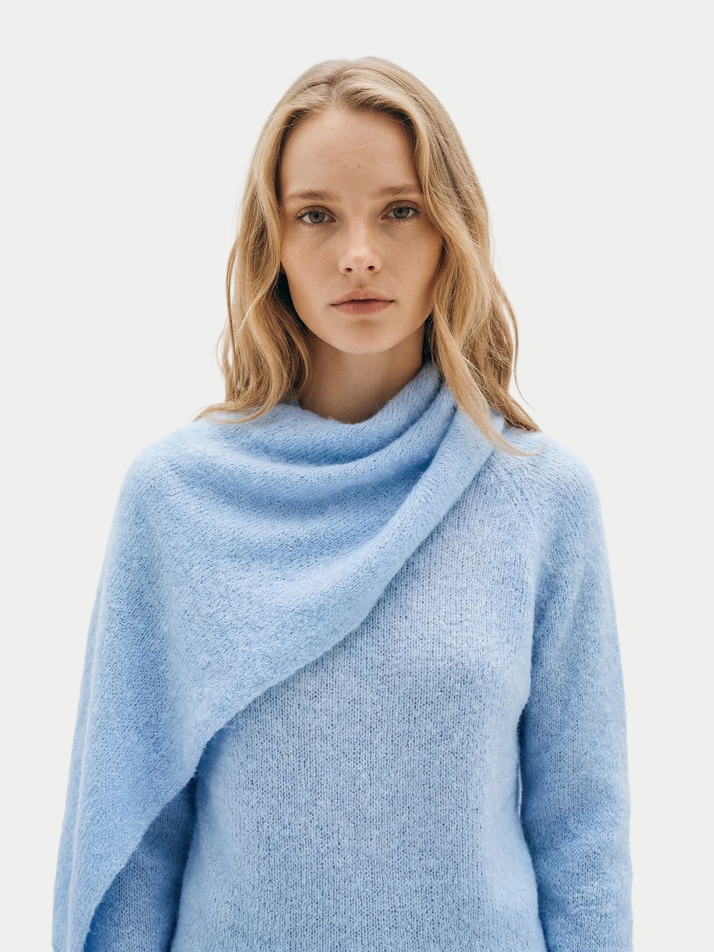 Vue de face d'une femme portant un pull bleu clair à manches longues et une écharpe intégrée, sur un fond gris clair. #color_bleu-clair