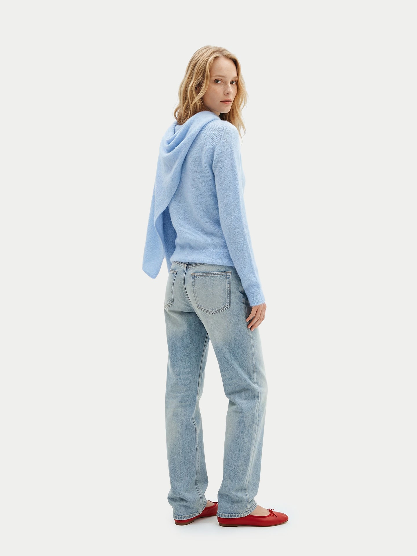 Vue de dos d'une femme debout portant un pull bleu clair à manches longues avec une écharpe intégrée, un jean bleu et des ballerines rouges sur un fond gris clair. #color_bleu-clair