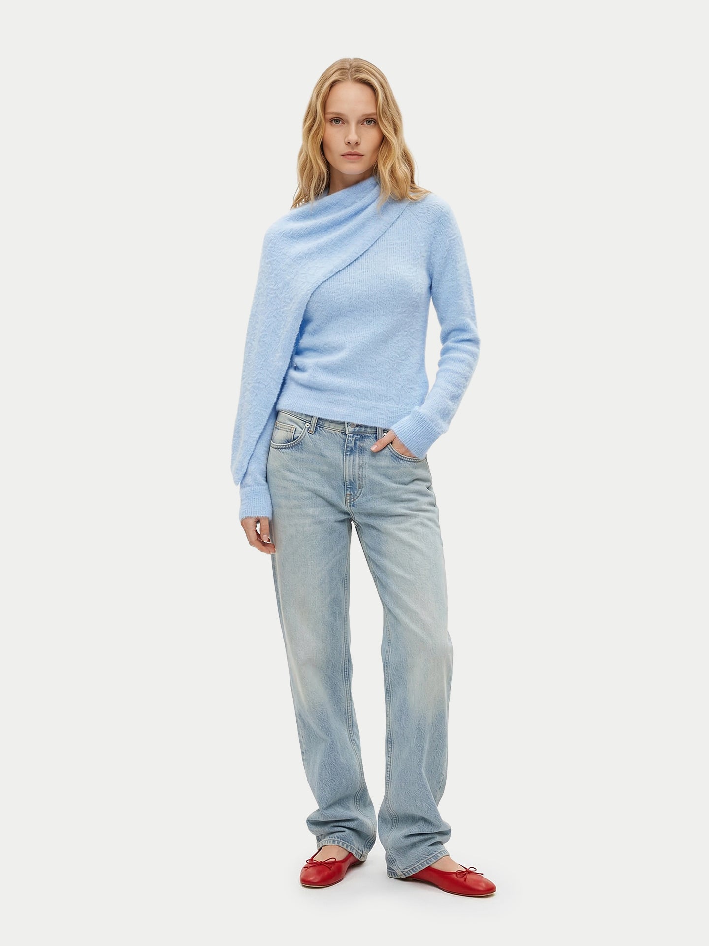 Vue de face d'une femme debout portant un pull bleu clair à manches longues avec une écharpe intégrée, un jean bleu et des ballerines rouges sur un fond gris clair. #color_bleu-clair