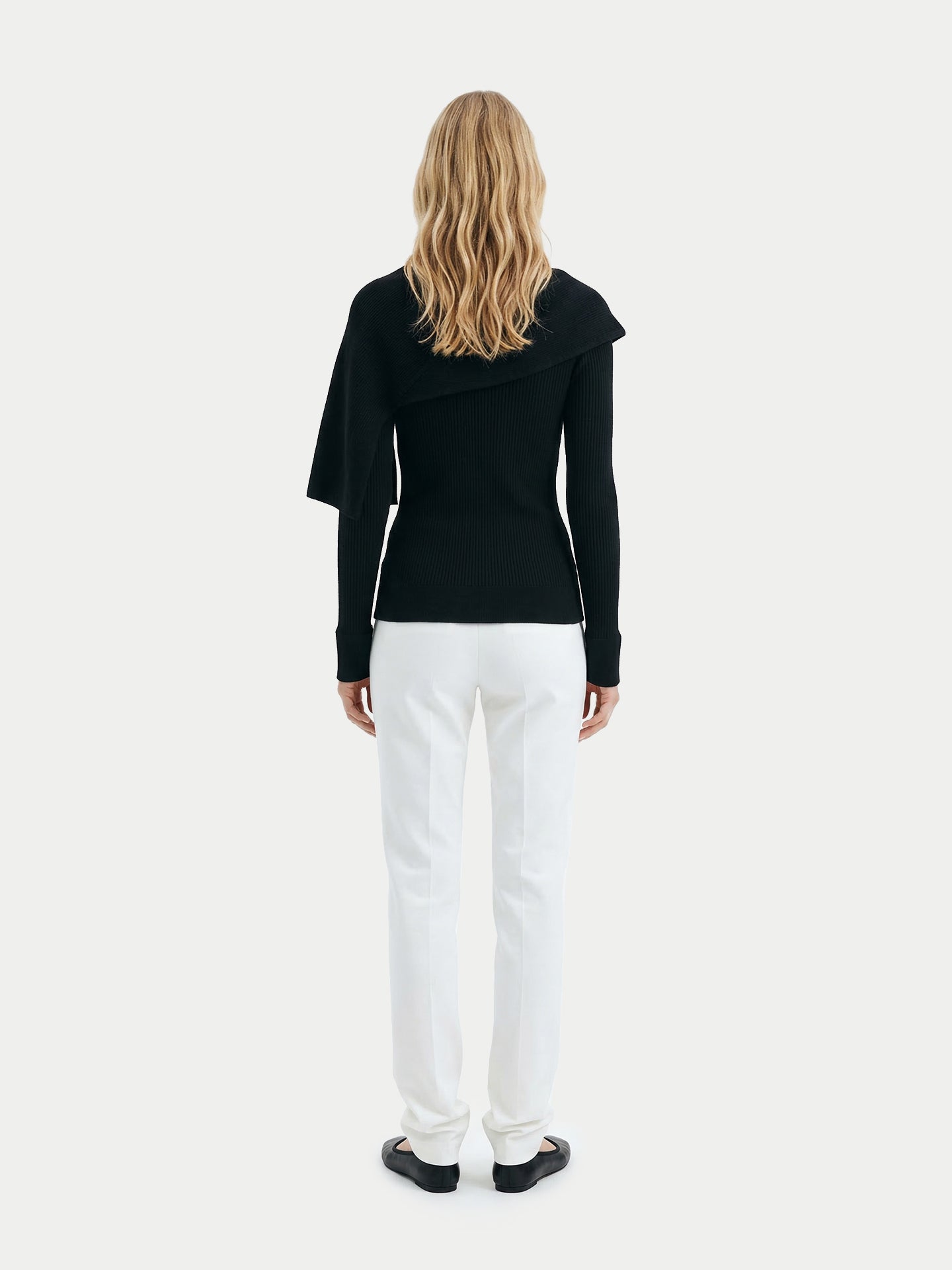 Vue de dos d'une femme portant un pull noir à manches longues avec une écharpe intégrée, un pantalon blanc et des ballerines noires sur un fond gris clair. #color_blanc
