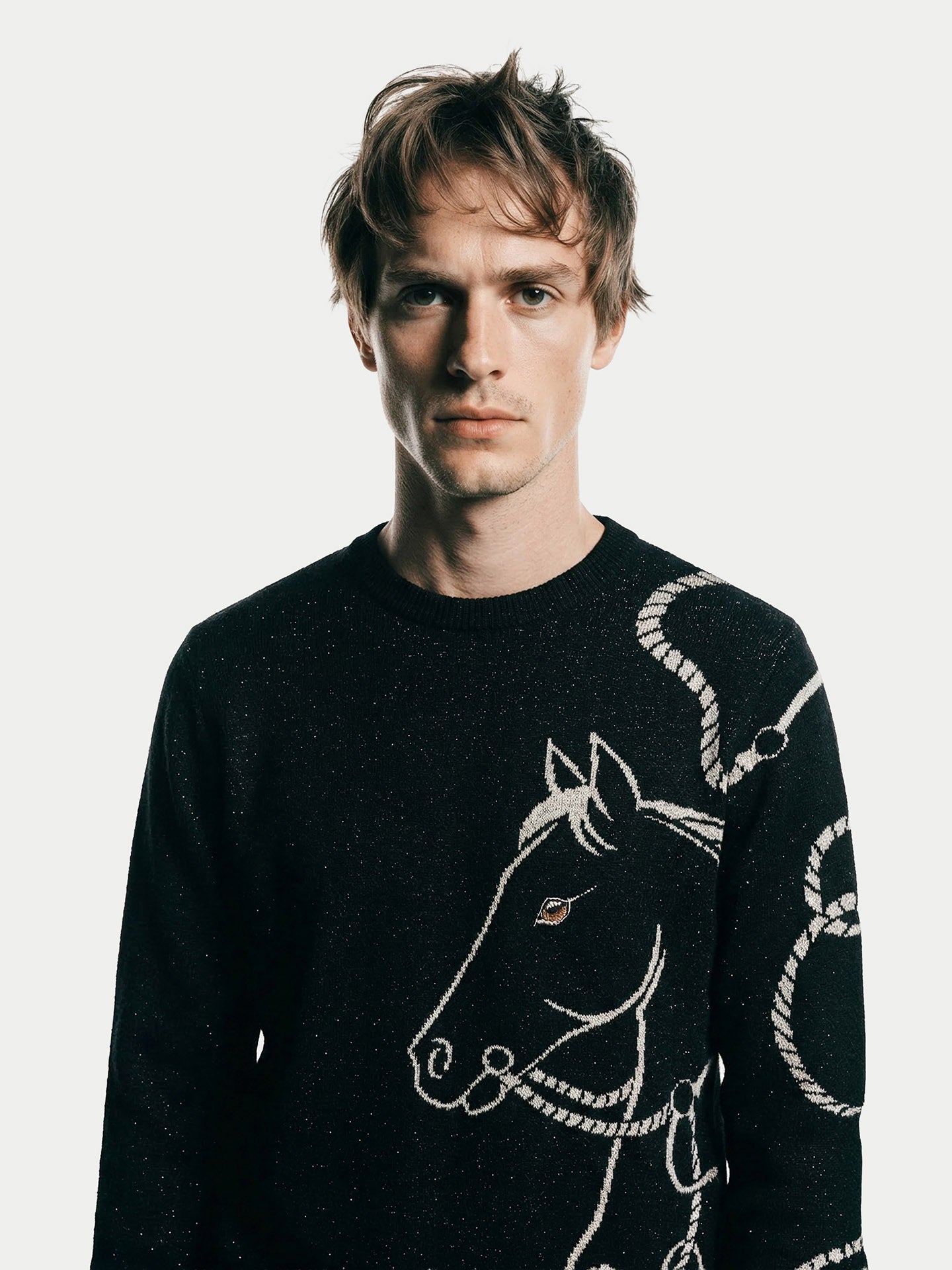 Portrait de face d'un homme aux cheveux châtains ébouriffés, portant le pull en cachemire noir avec le motif blanc de tête de cheval et de cordes. Il regarde directement l'objectif sur un fond blanc épuré. #color_noir
