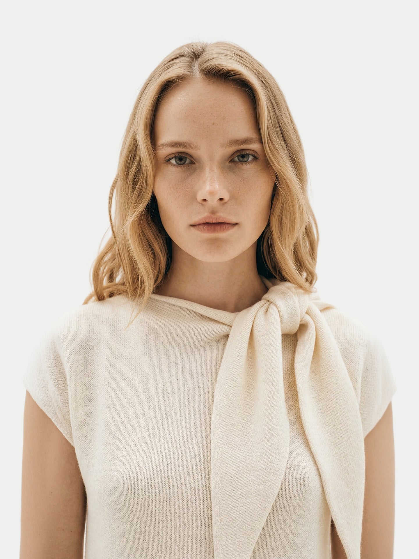 Gros plan de face d'une femme portant une robe blanche à manches courtes arrivant sous le genou, avec un foulard noué, sur un fond gris clair. #color_blanc