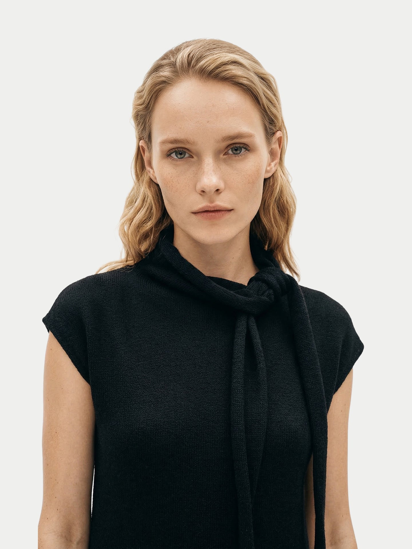 Gros plan de face d'une femme portant une robe noire à manches courtes arrivant sous le genou, avec un foulard noué, sur un fond gris clair. #color_noir