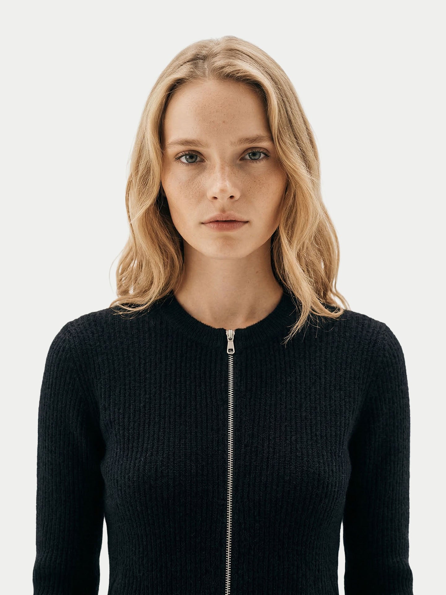 Gros plan de face d'une femme portant un cardigan noir à col rond, zippé et côtelé, sur un fond gris clair. #color_noir